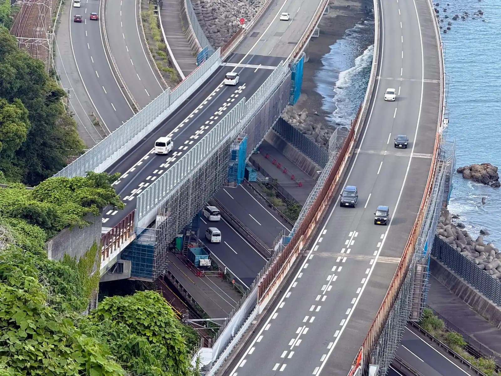 This is what you climb the pass to see. The Tokaido Main Line runs tight against the cliff, National Route 1 next to it, and the Tomei Expressway out over the water — all of it packed into one narrow strip of coast.