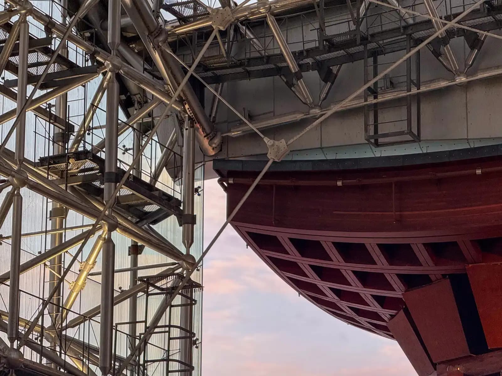 Where the wooden roof of the Tsuzumi Gate meets the steel frame and glass of the Motenashi Dome, with a pink evening sky between them.