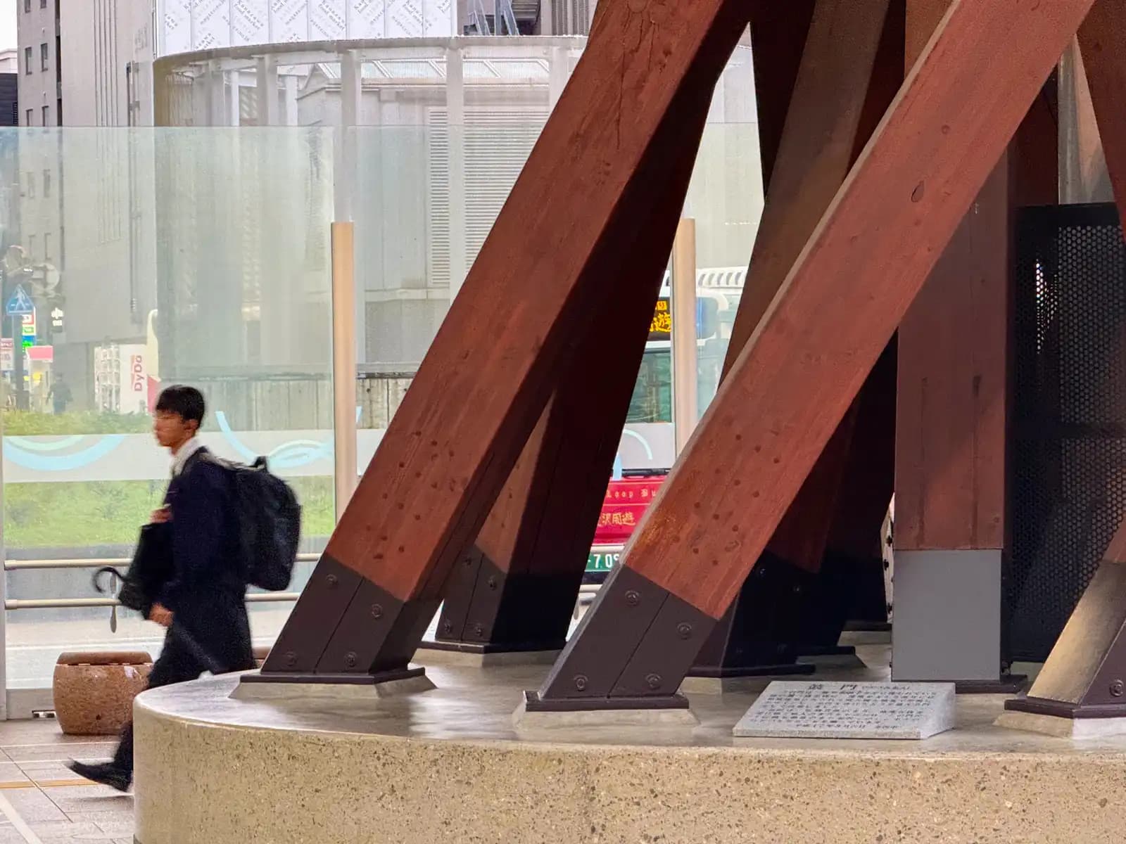 A high school student walks past the base of the Tsuzumi Gate, where the wooden pillars are bolted to steel footings on a concrete platform.