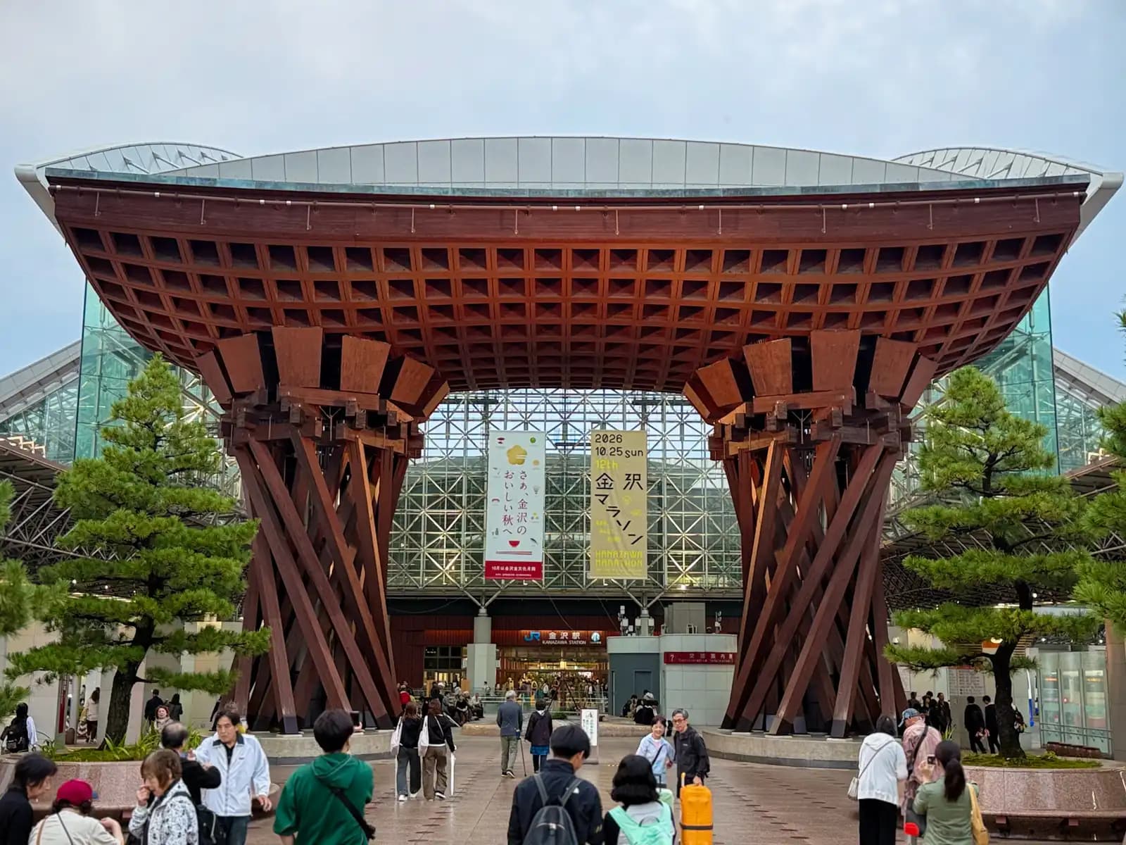 The Tsuzumi Gate from the front, with pine trees on both sides and the Motenashi Dome behind it. People walk to and from the station entrance under the gate.
