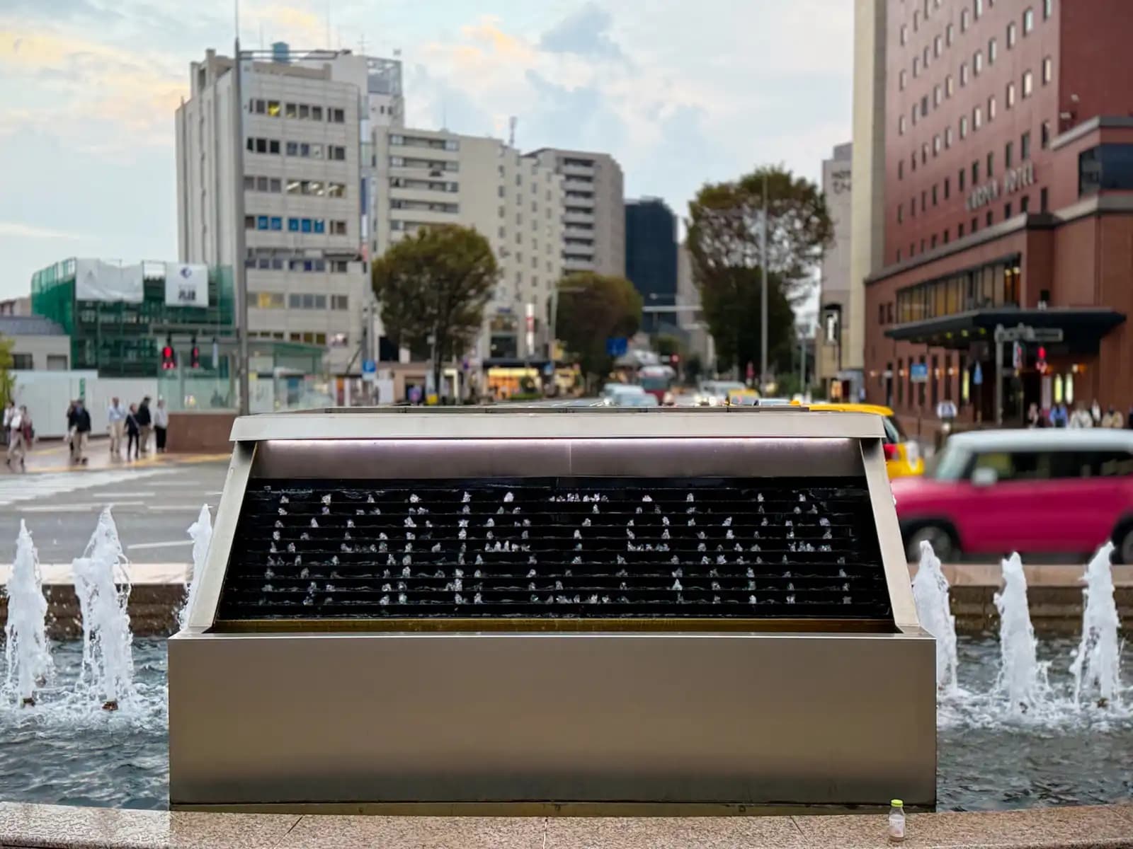 The water clock fountain in front of the station's east exit. Water drops fall in patterns to show the time and short messages, with the street and hotels behind.