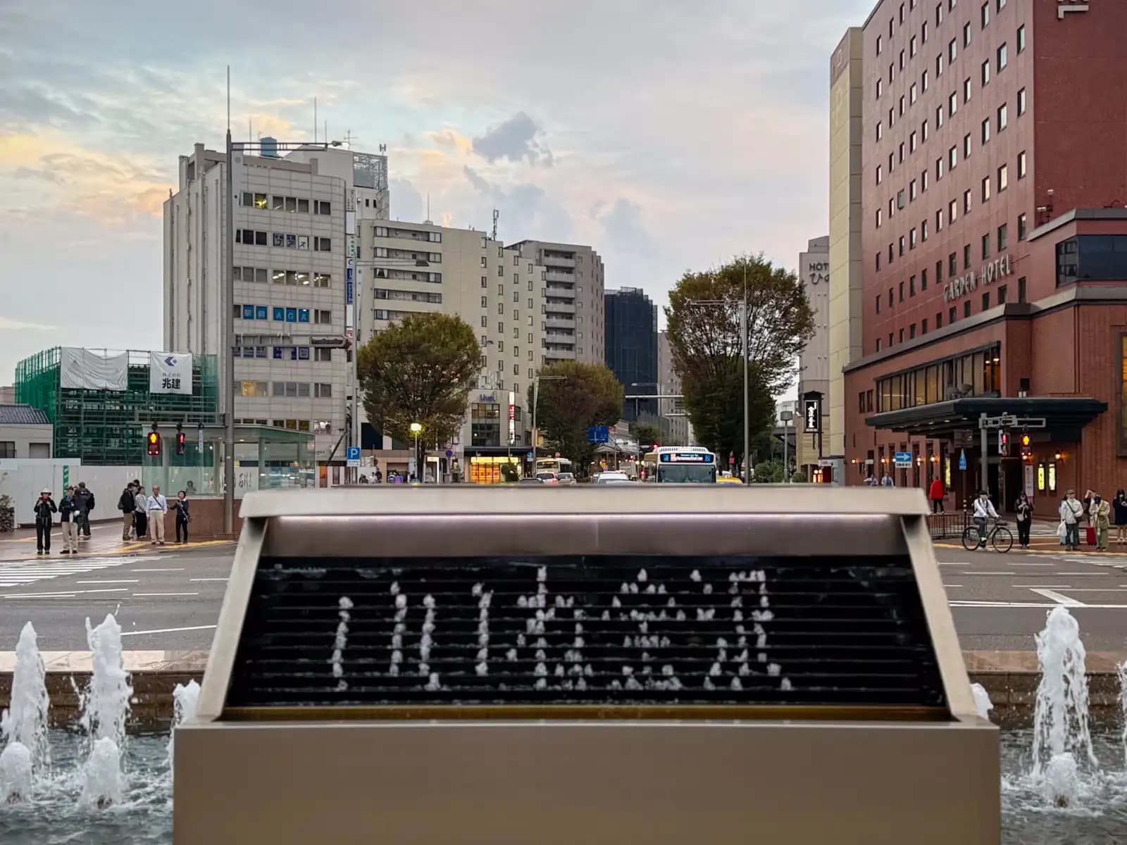 The water clock fountain spelling out いいね金沢 in falling water drops. It roughly means "Kanazawa is great" — いいね is the everyday Japanese word for "nice" or "good," the same word used for a "like" on social media. The main road leading away from the station and a hotel are behind.