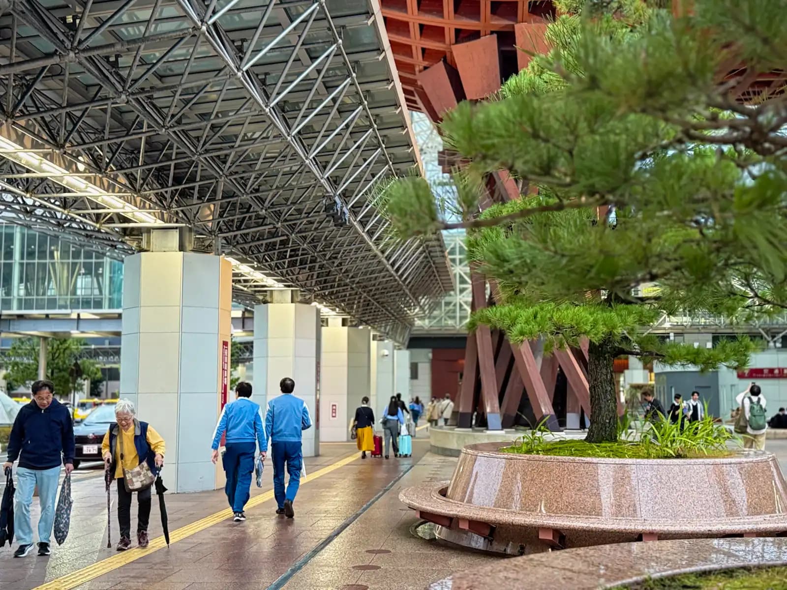 People walking along the covered walkway outside the station. A pine tree grows from a round stone planter with a bench built around it, and the Tsuzumi Gate is behind.