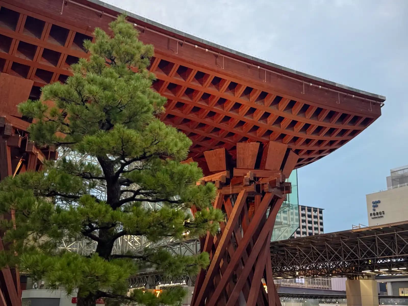 A pine tree in front of the Tsuzumi Gate's wooden roof. The Forus shopping mall is on the right.