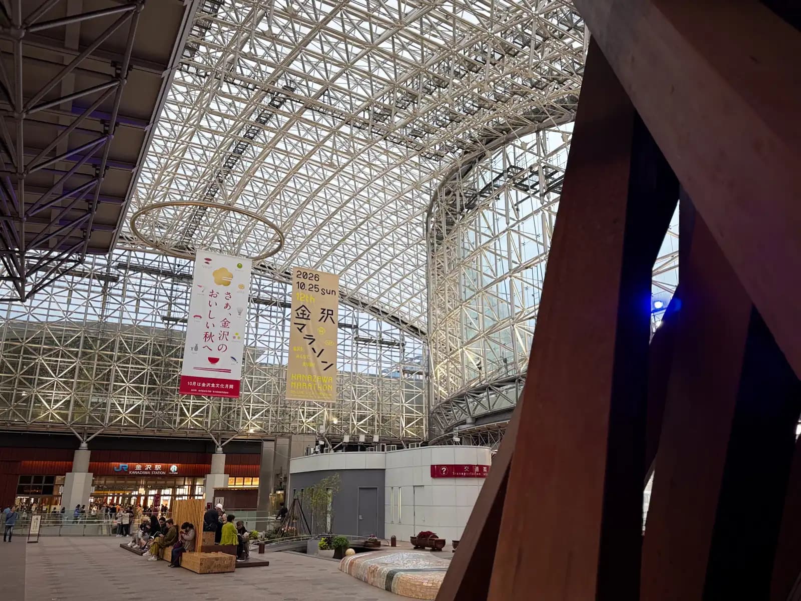 The Motenashi Dome seen from beside one of the Tsuzumi Gate's pillars. People sit on wooden benches in the plaza in front of the station entrance.
