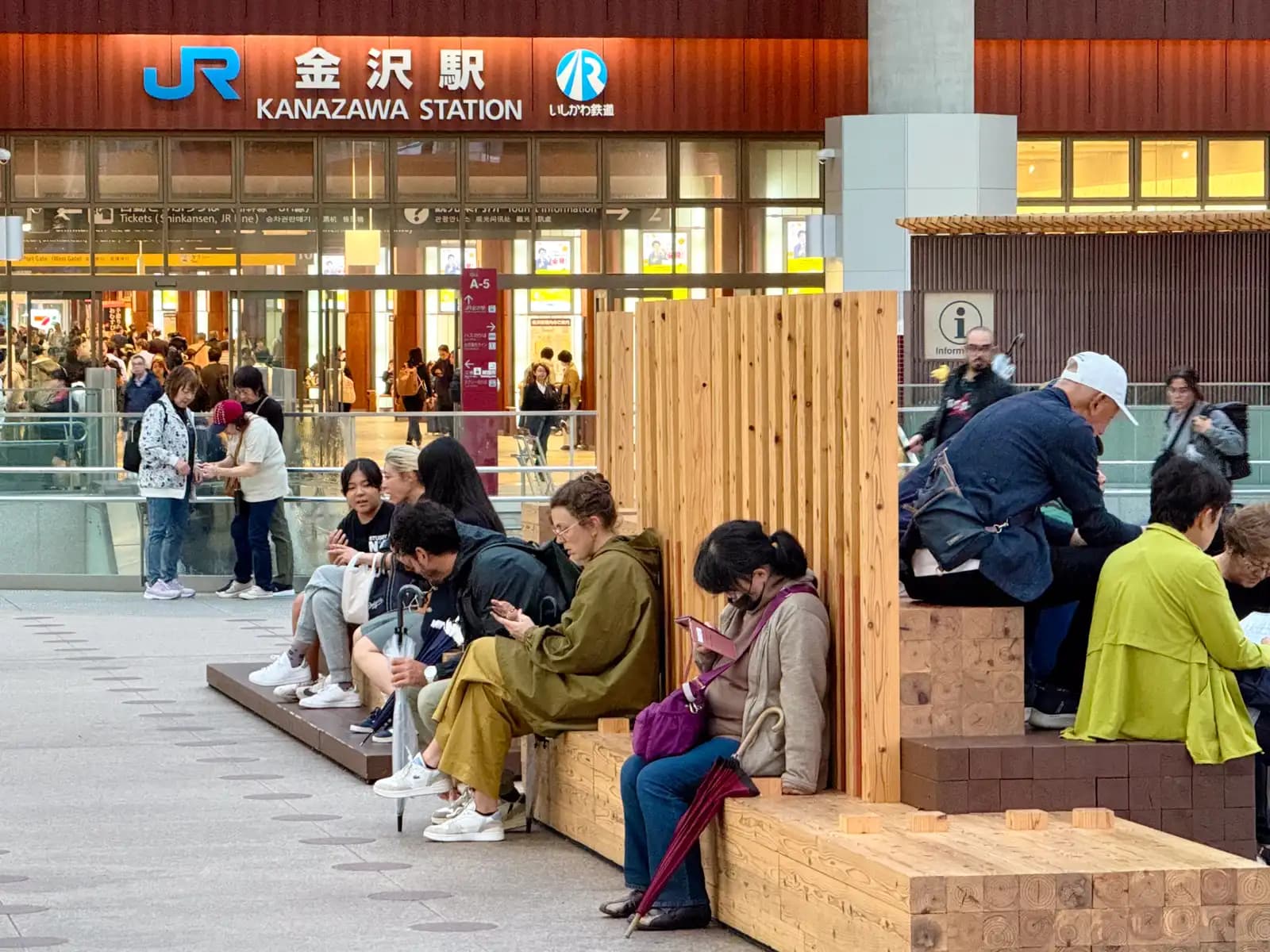 People resting on wooden benches in front of the JR Kanazawa Station entrance. The sign above shows both JR and the IR Ishikawa Railway, which share the station.
