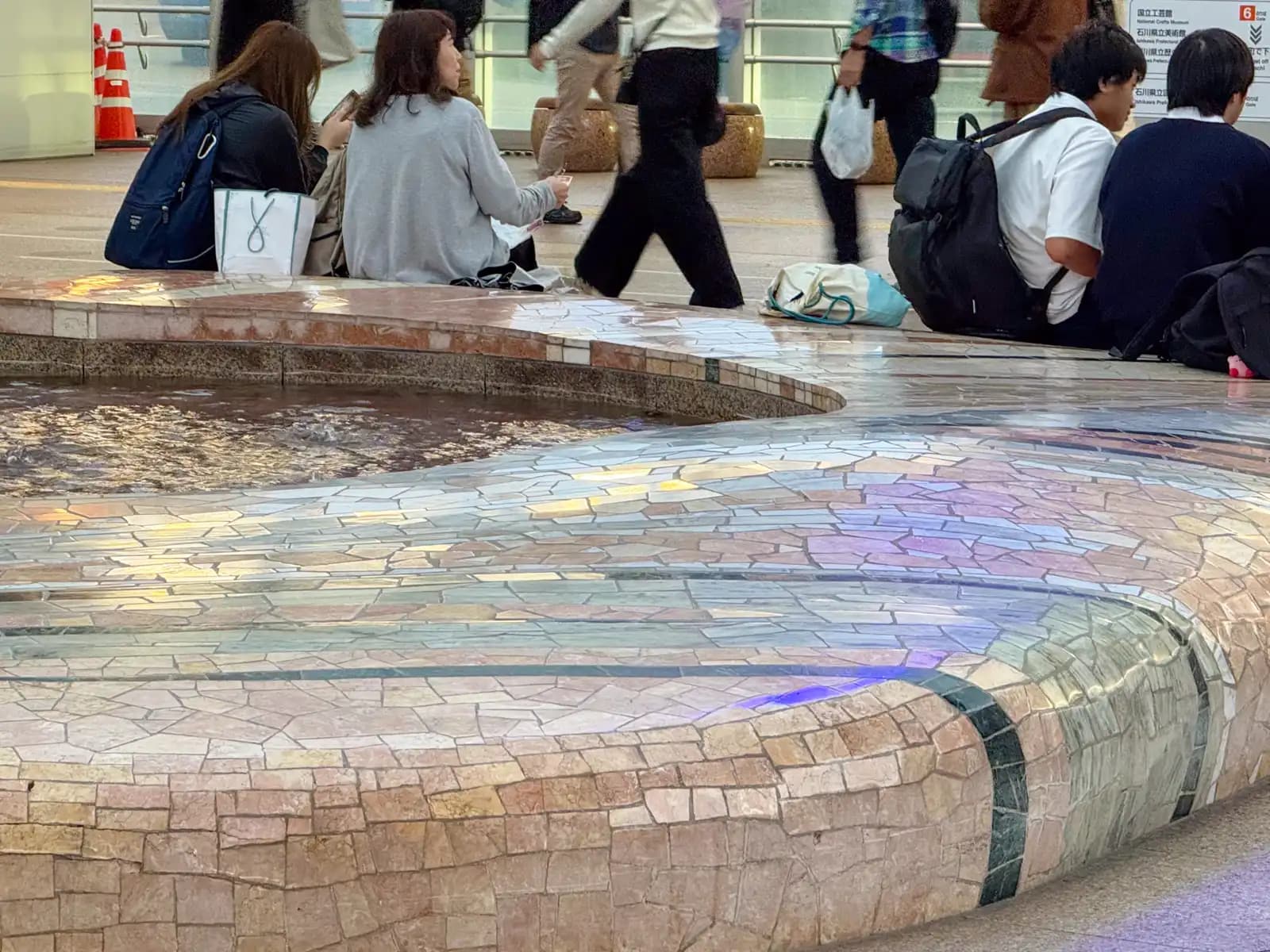 The curved edge of a small fountain in the station plaza, covered in pieces of colored stone. People sit on the edge to rest.