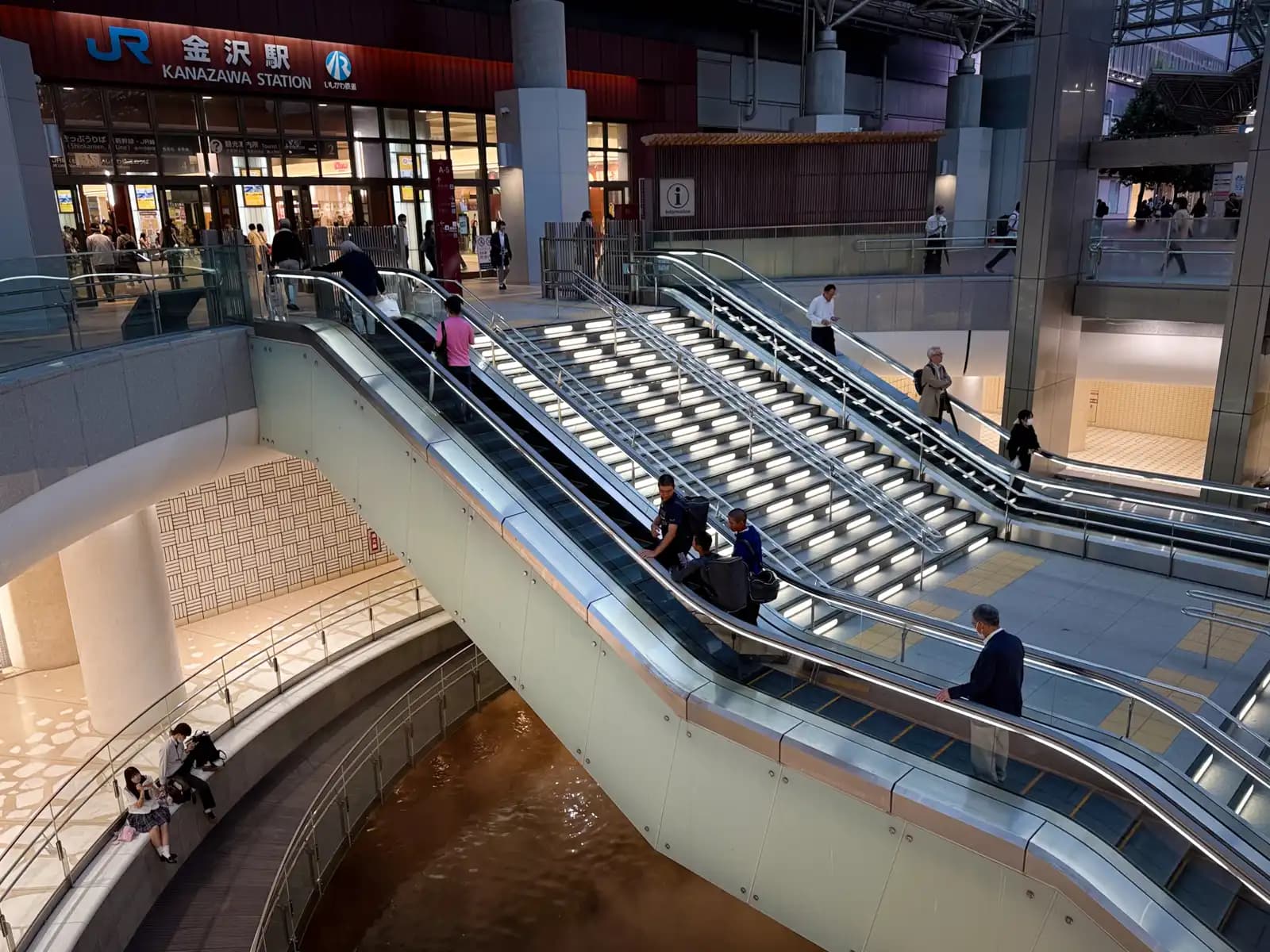 Escalators and stairs leading from the JR Kanazawa Station entrance down to the underground level, with lights set into each step.
