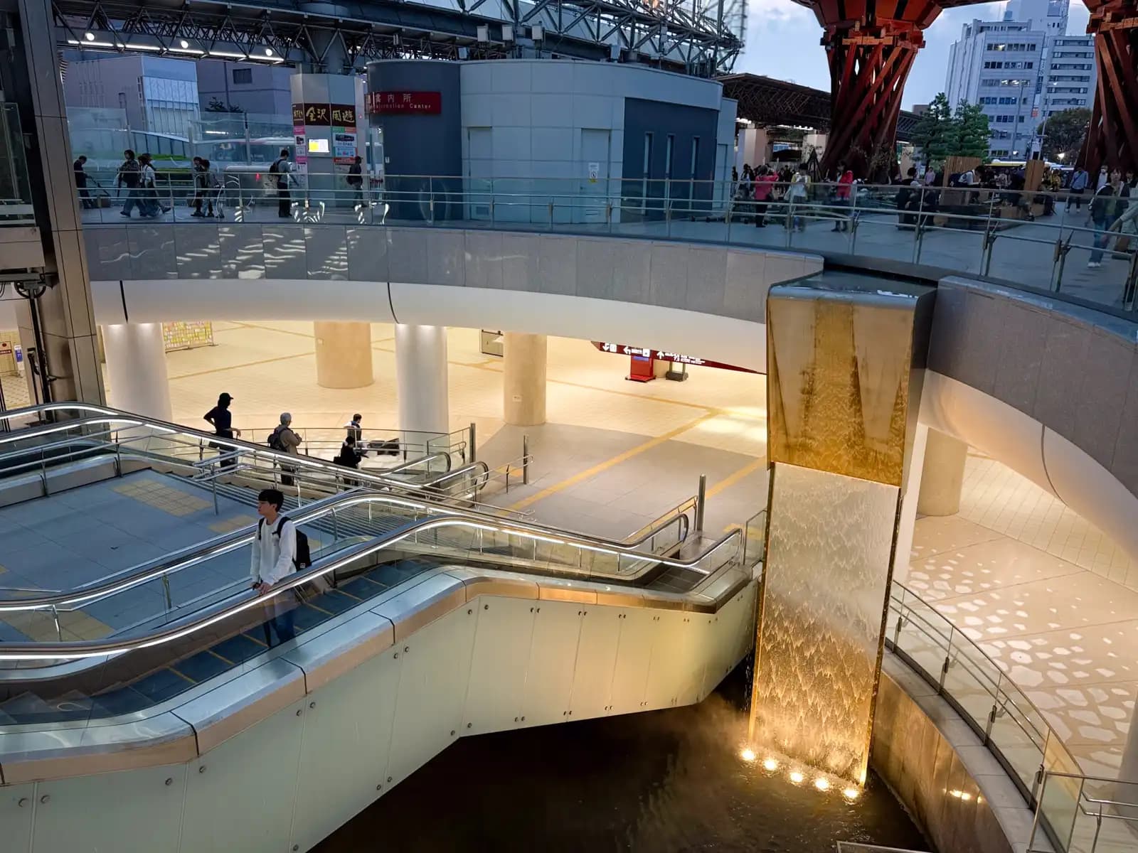 The sunken plaza below the station's east exit. Water runs down a lit wall next to the escalators, and the Tsuzumi Gate is visible at street level above.