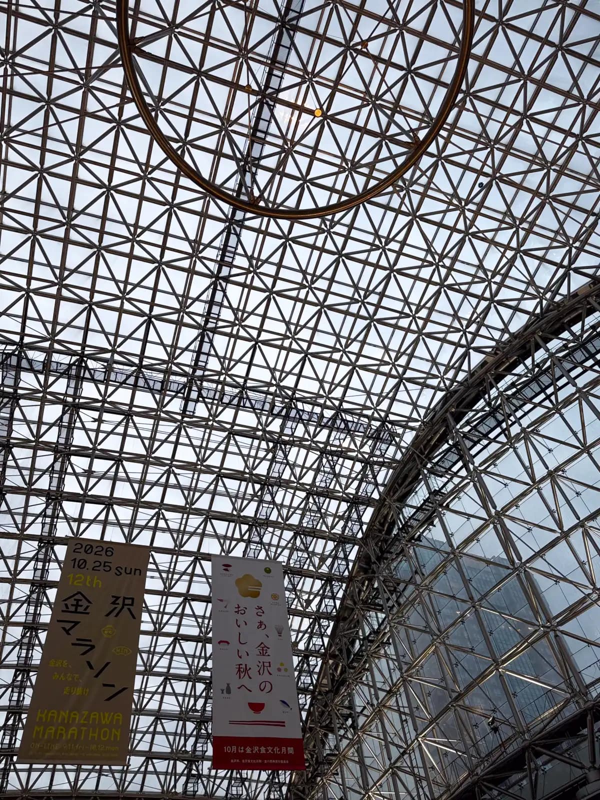 Looking up at the glass roof of the Motenashi Dome, with a large metal ring hanging from the center and banners for the Kanazawa Marathon and autumn food events.