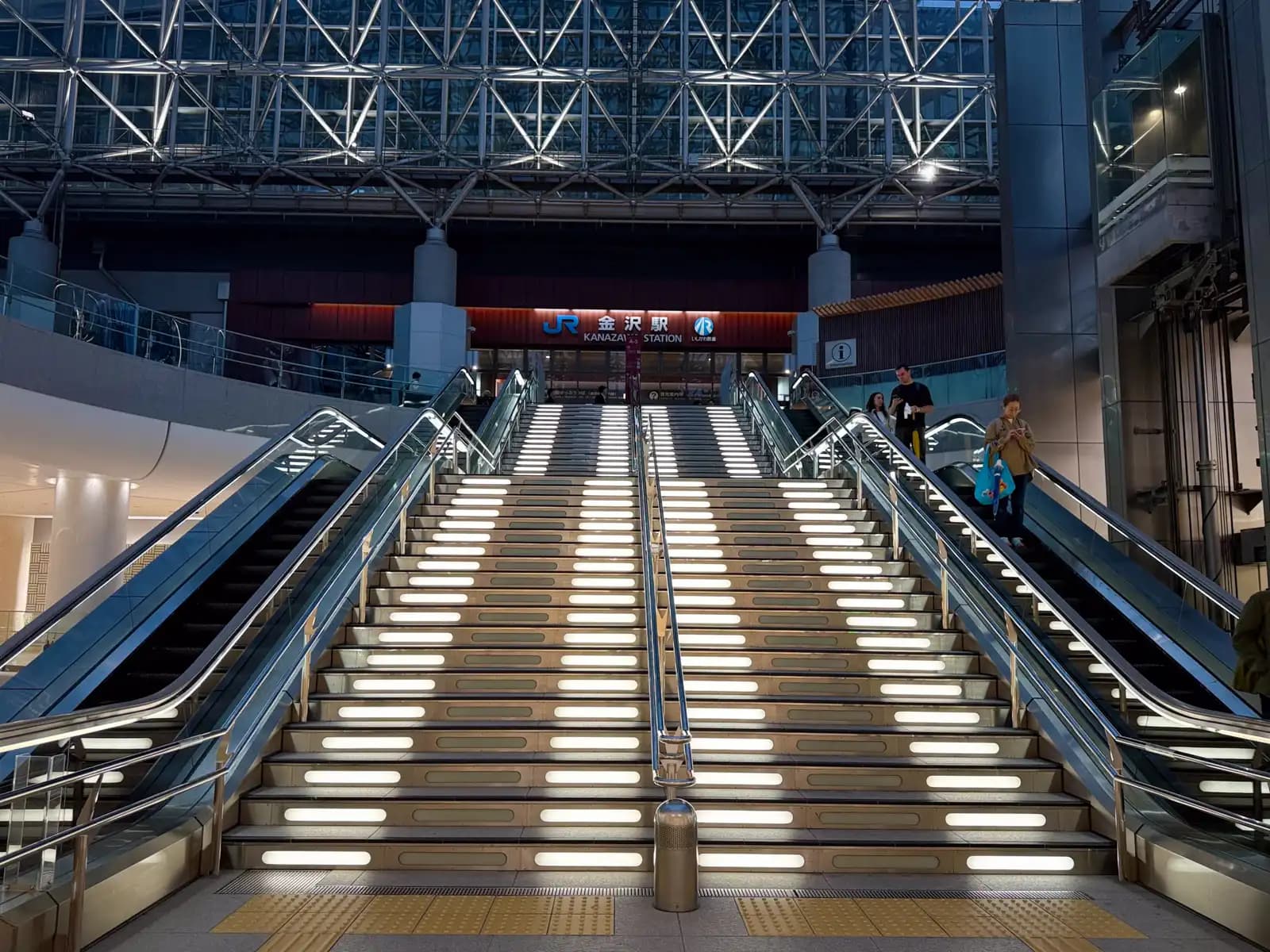 Lit stairs with escalators on both sides, leading up from the underground plaza to the JR Kanazawa Station entrance.