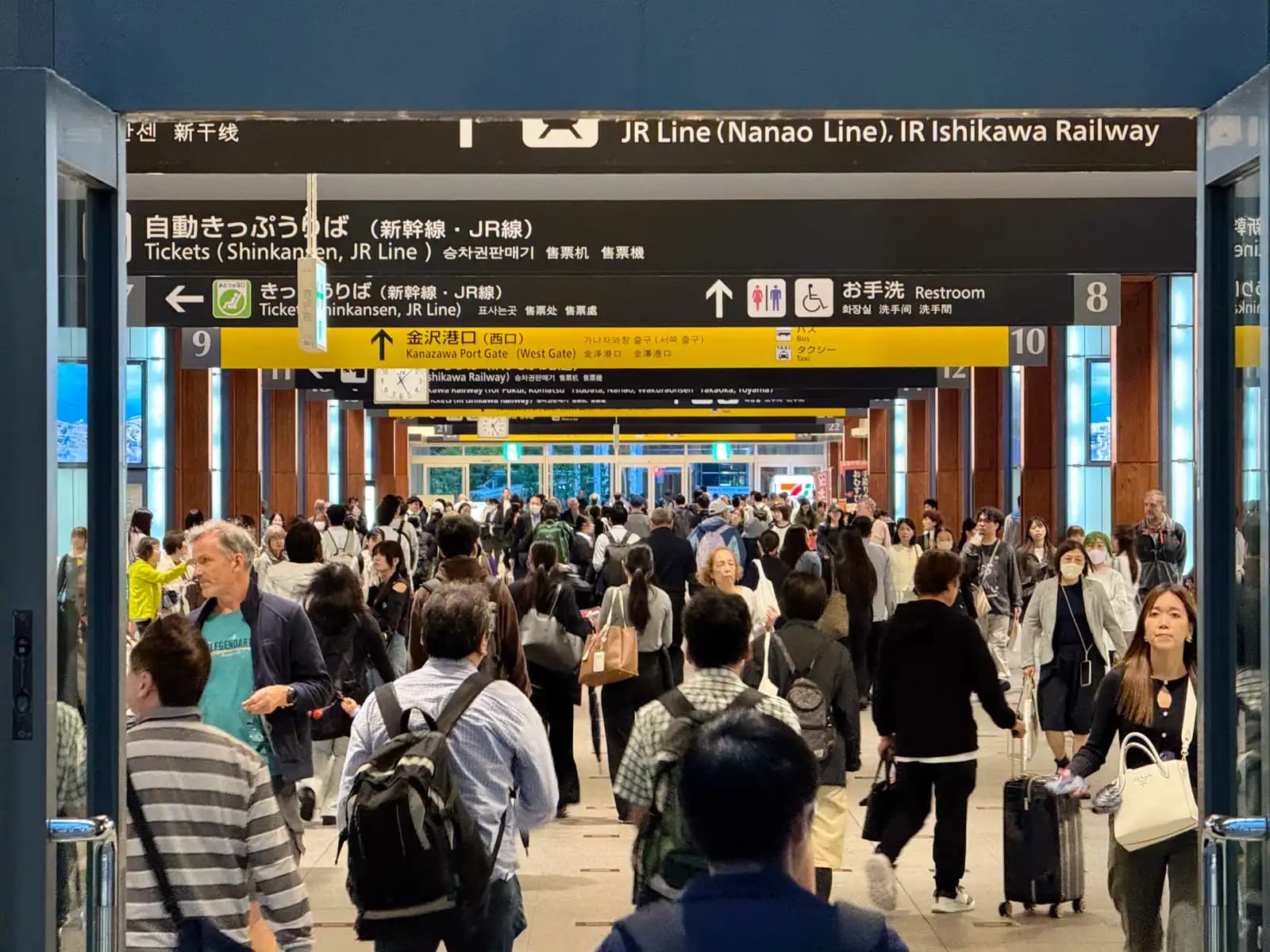 A busy concourse inside Kanazawa Station. Signs point to the ticket machines, the restrooms, the JR and IR Ishikawa Railway lines, and the West Gate.