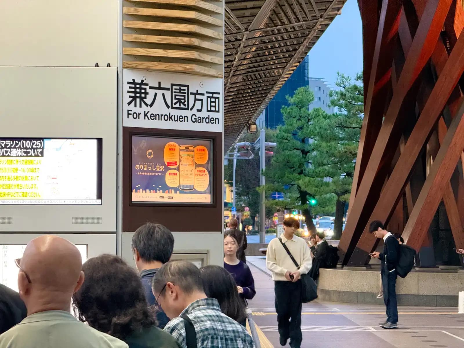 People lined up at the bus stop for Kenrokuen Garden, next to the Tsuzumi Gate. Kenrokuen is counted as one of the three great gardens of Japan. The screen on the left gives notice of bus delays on the day of the Kanazawa Marathon.