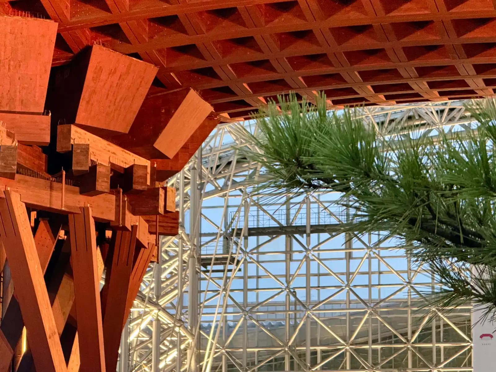 Close-up of the wooden joinery at the top of a Tsuzumi Gate pillar, with a pine branch and the steel frame of the Motenashi Dome behind.