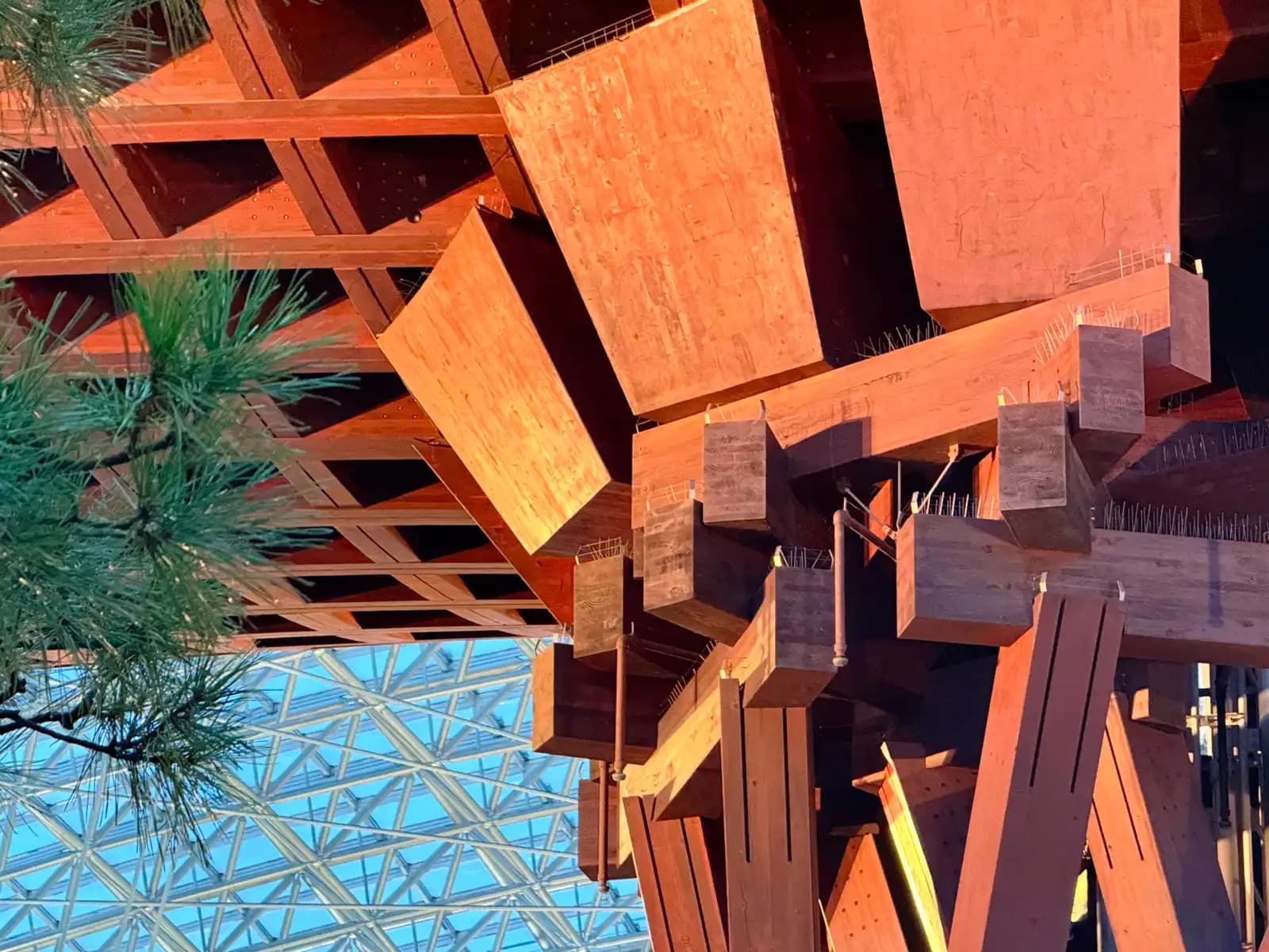 Looking straight up at the wooden blocks and beams where a Tsuzumi Gate pillar meets the roof, with a pine branch and the glass of the Motenashi Dome in the corner.