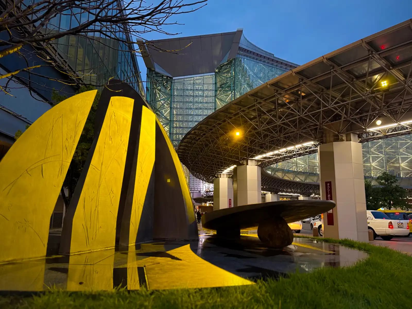 A sculpture lit in yellow and a stone bench on the lawn by the station's taxi area in the evening. The station building and its glass walls are behind.