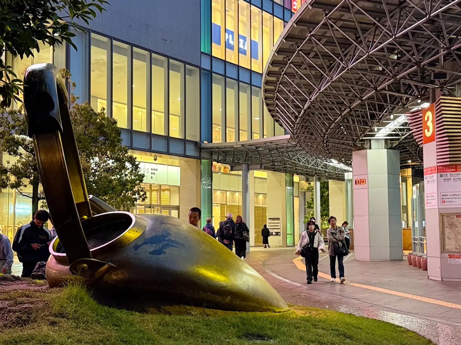 A large bronze sculpture of a kettle lying on its side on the lawn next to bus stop 3, in front of the shopping buildings by the station's east exit.