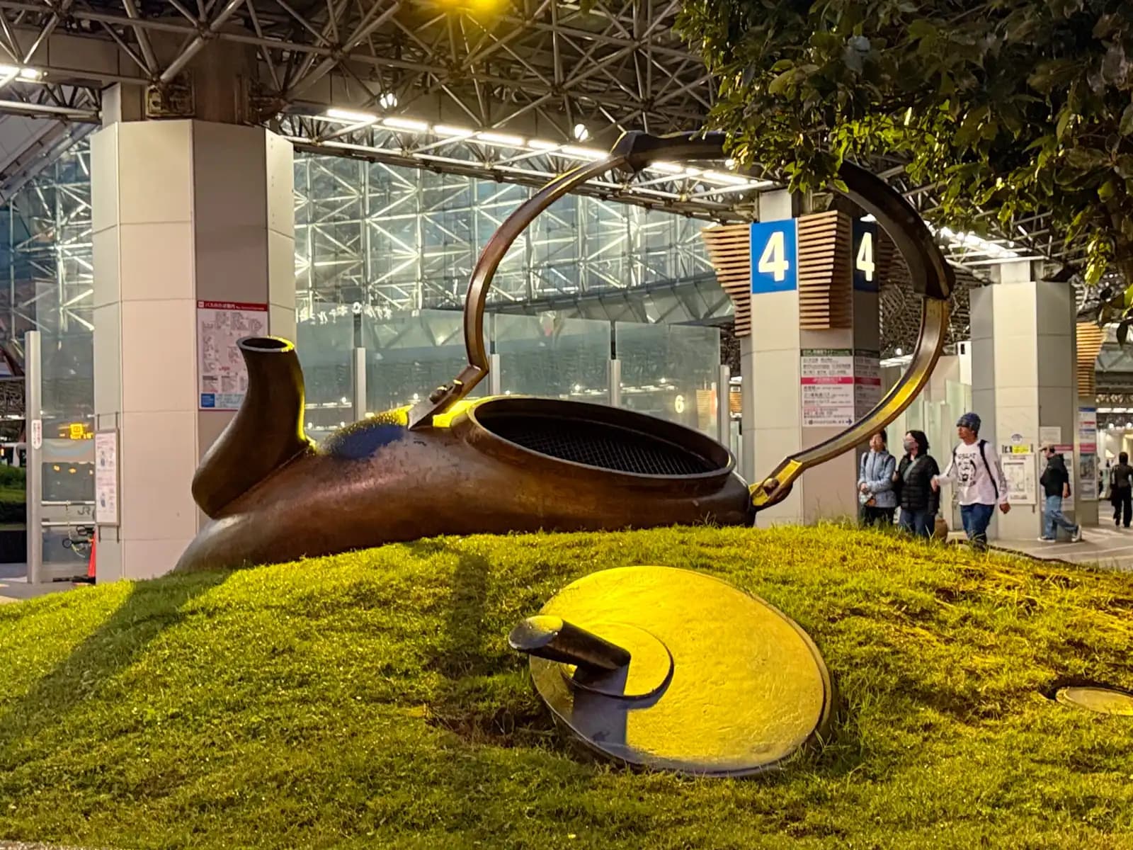 The bronze kettle sculpture with its lid lying on the grass beside it. Bus stop 4 is behind.