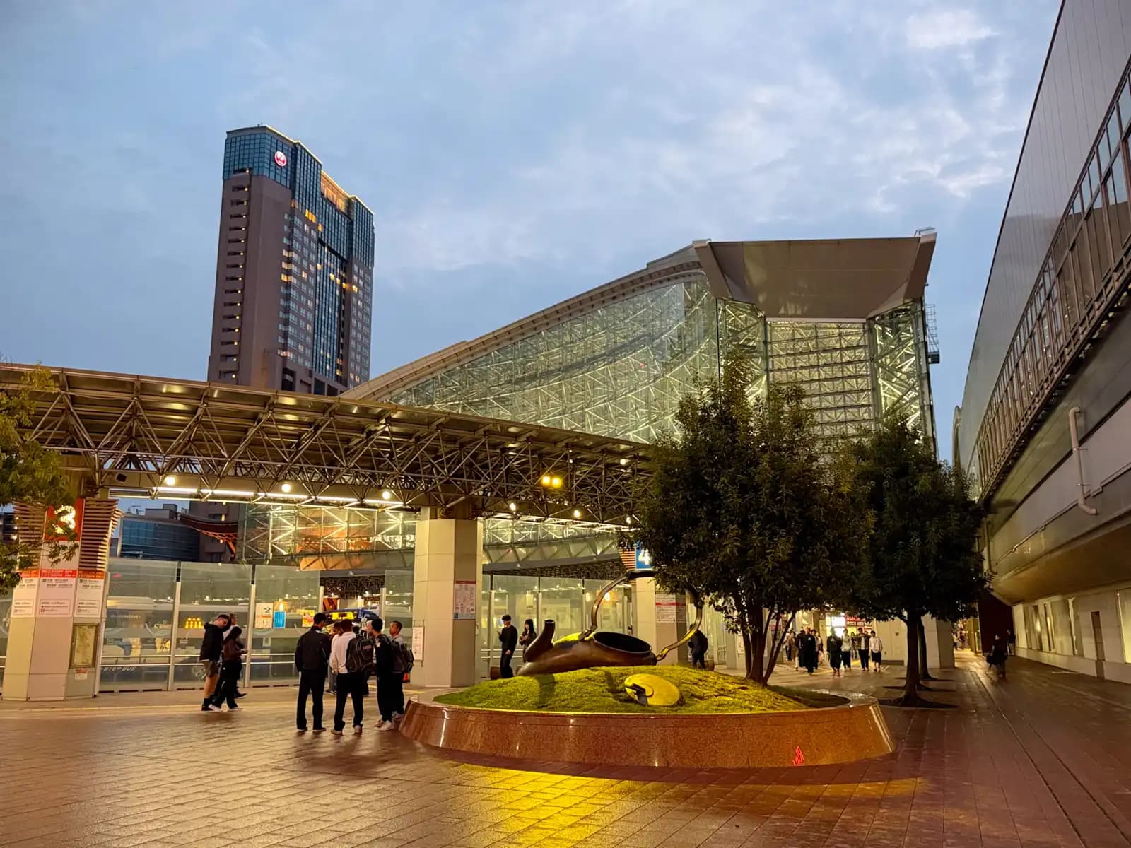 The kettle sculpture on its raised lawn in the evening, with the Motenashi Dome behind it and the ANA Crowne Plaza hotel tower on the left.