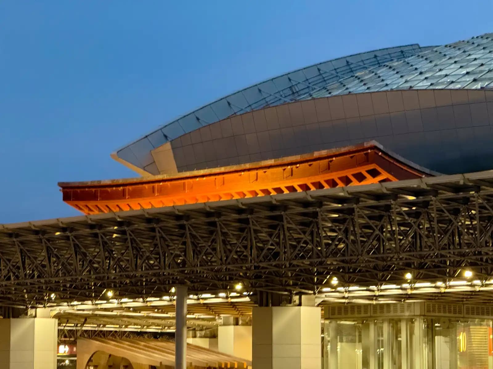 The roof of the Tsuzumi Gate lit in orange at dusk, below the edge of the Motenashi Dome. The covered walkway around the bus terminal is in front.