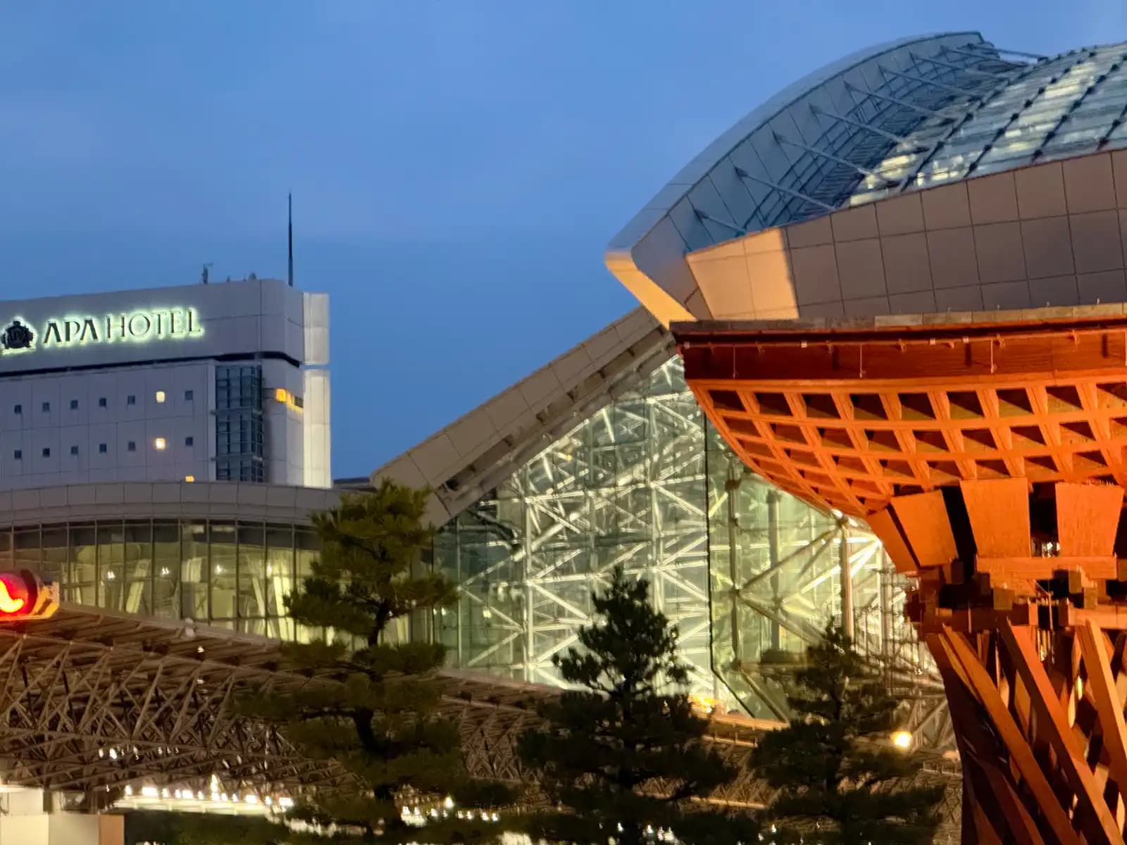 The Tsuzumi Gate and the lit Motenashi Dome at dusk, with pine trees in front and the APA Hotel on the left.