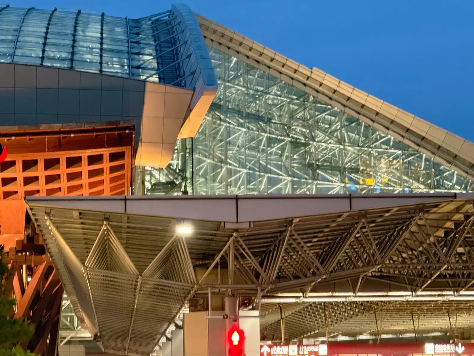 The glass side of the Motenashi Dome lit up at dusk, above the steel roof of the covered walkway. Part of the Tsuzumi Gate is on the left.