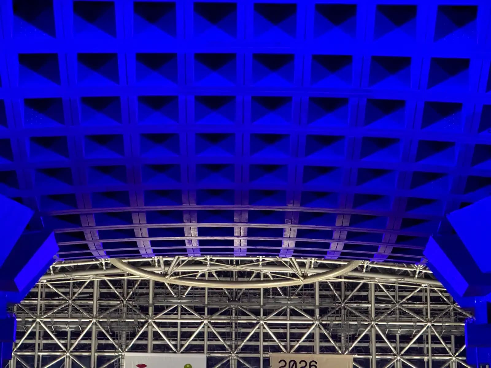 The underside of the Tsuzumi Gate's roof lit in blue at night, with the steel frame of the Motenashi Dome and its metal ring behind.