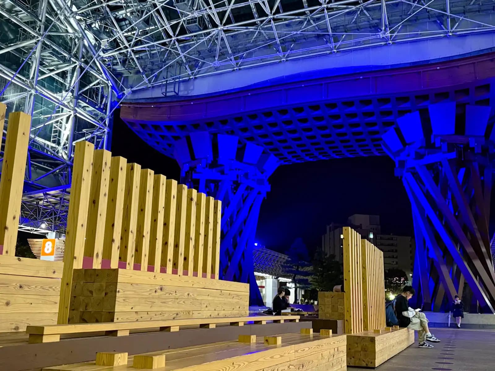 Wooden benches under the Motenashi Dome at night, with the Tsuzumi Gate lit in blue behind them. A few people sit and look at their phones.