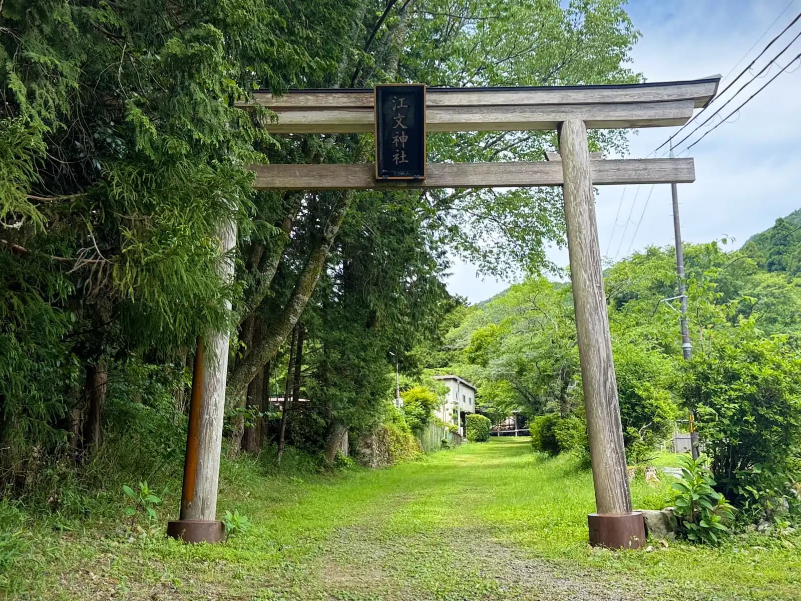 The weathered wooden torii at the entrance to Ebumi Shrine, its plaque reading 江文神社. A grass-covered path leads in from the edge of Ohara toward the forested foot of Mt. Konpira.