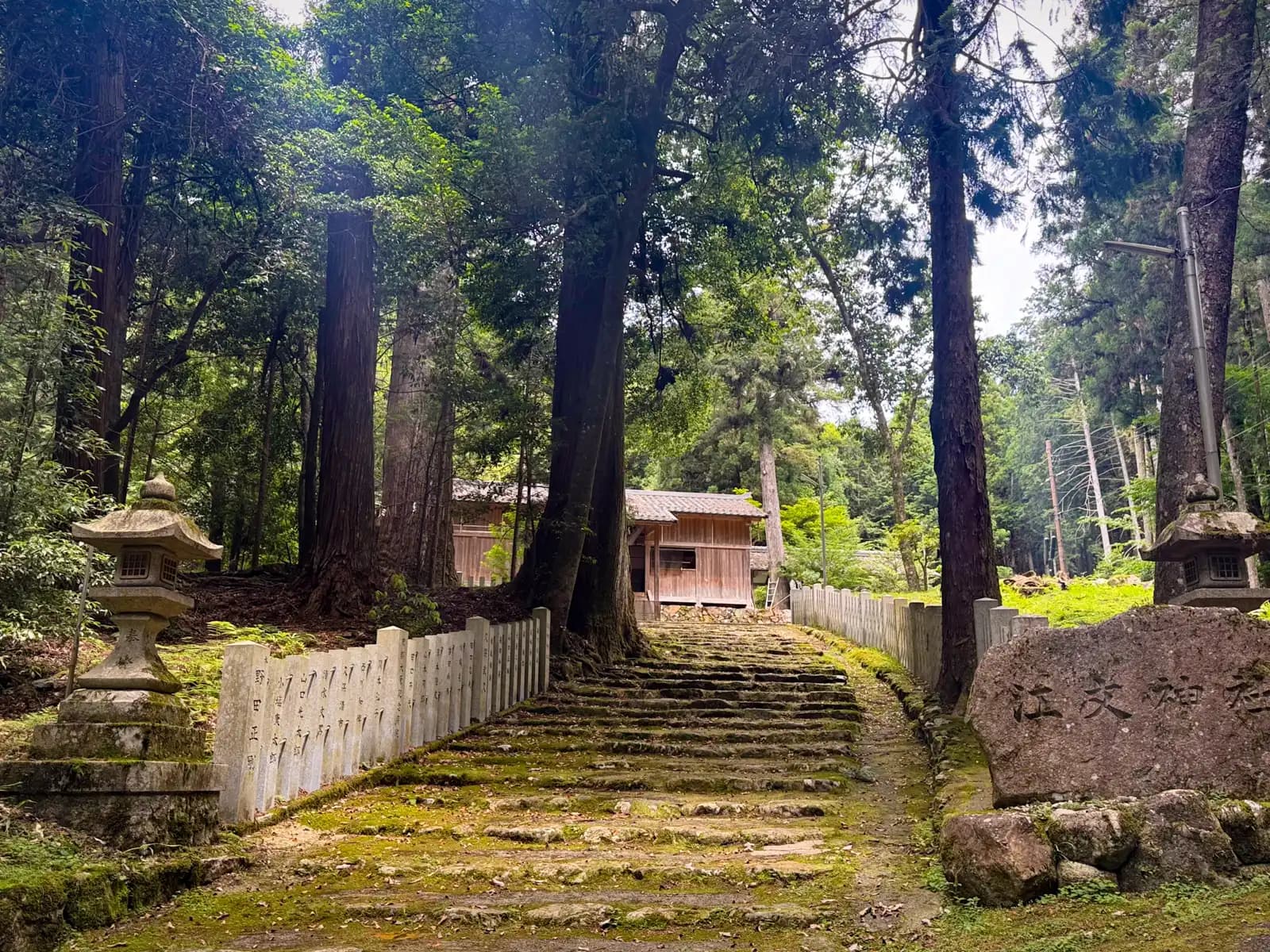 Moss-covered stone steps climb between stone lanterns and a low inscribed balustrade. The boulder at right is carved 江文神社; the shrine halls wait among the cedars above.