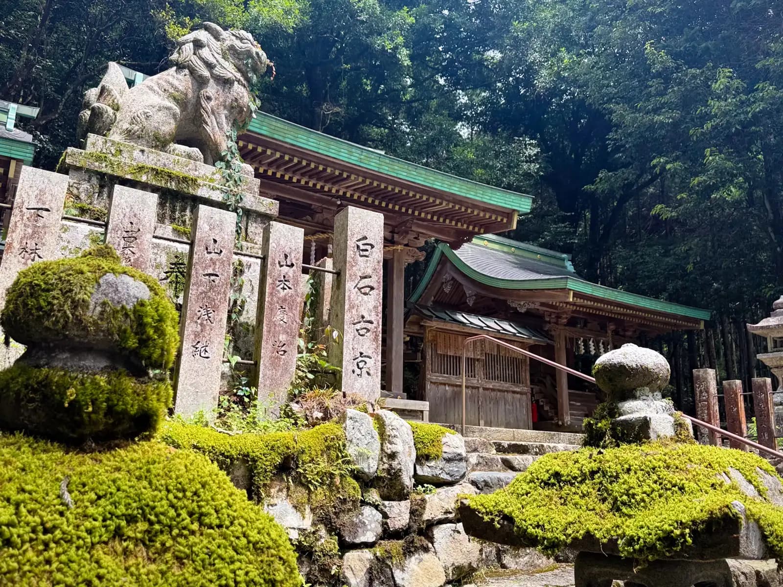 A moss-clad komainu perched above stone pillars inscribed with donors' names, with a weathered stone lantern and a green-roofed side hall behind.