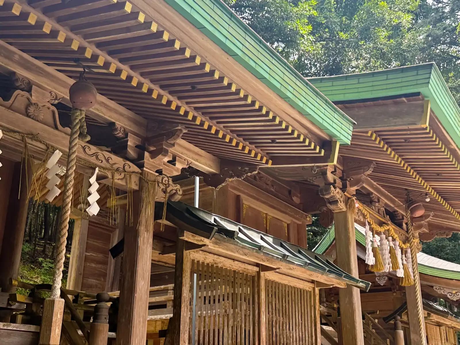 Carved brackets and gold-tipped rafters beneath the shine's roofs, where a suzu bell hangs from its braided rope and paper shide. The main hall meets a side hall in a run of joinery.