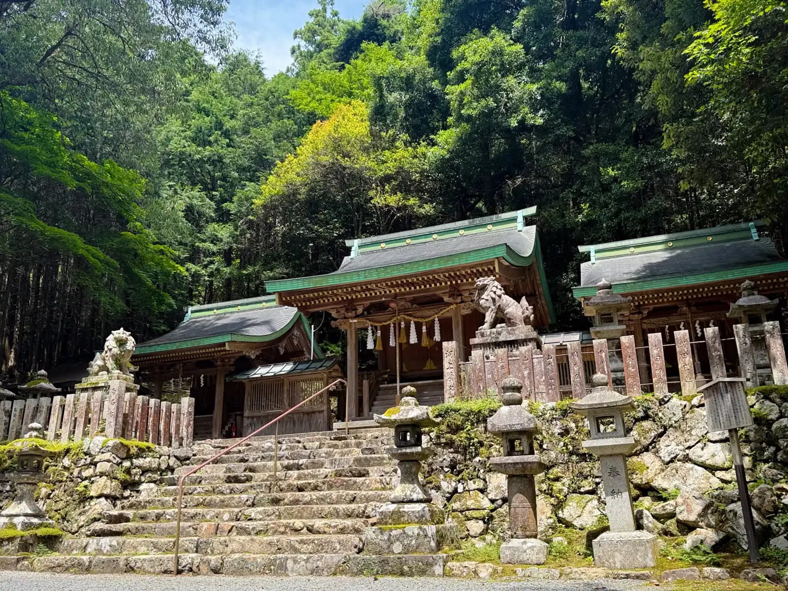 The shrine in full sun: the three halls with their copper-green roofs, a komainu at the steps, and rows of stone lanterns and donor pillars along the masonry, below the forested mountainside.