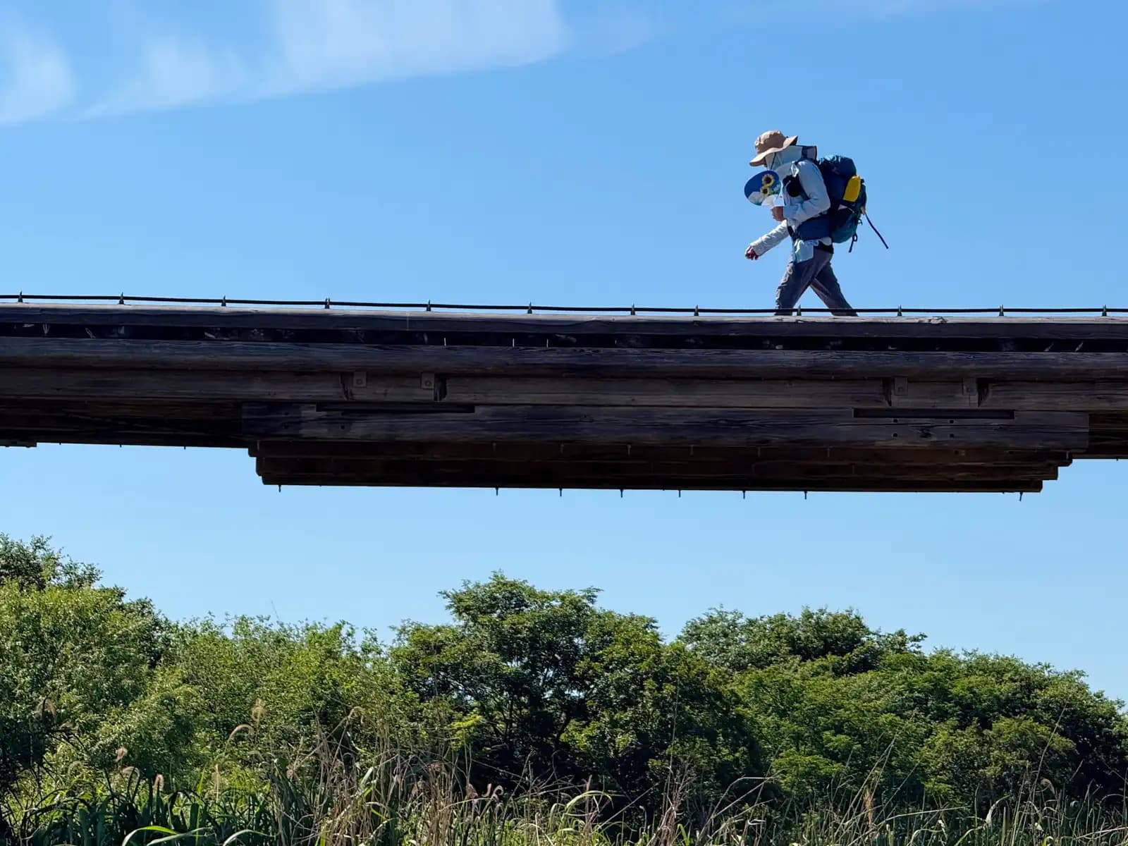 A backpacker walks the railless deck against an empty summer sky. Nothing between the planks and the drop but sure footing.