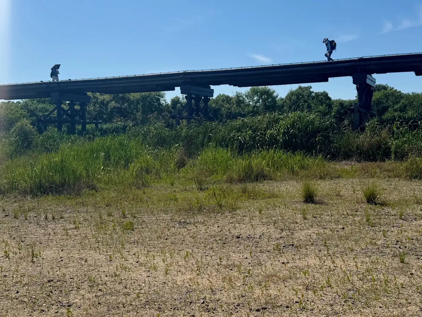 Two walkers, one under a parasol, make their way along the deck under a broad sky, seen across the dry riverbed.