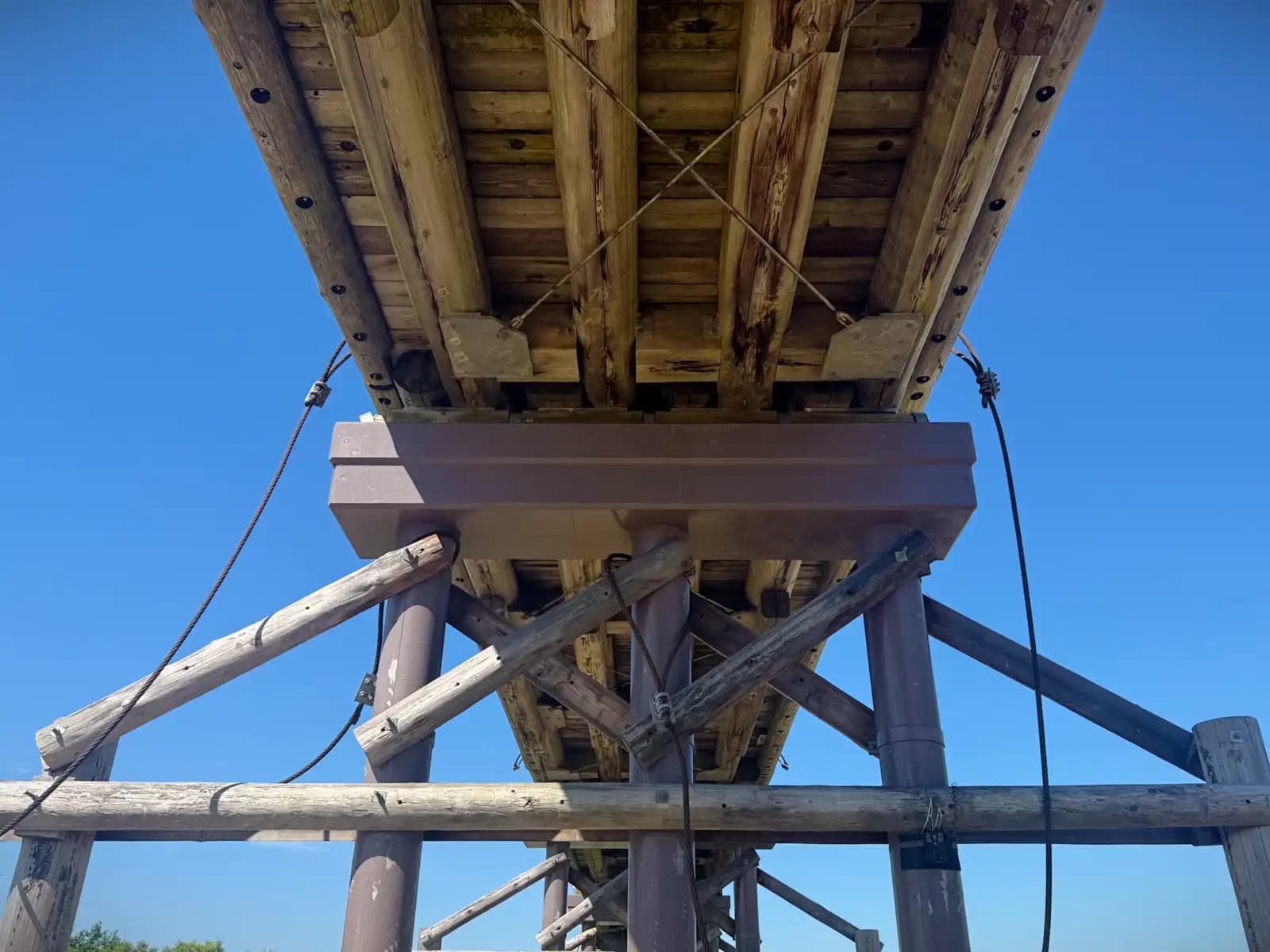 Straight up into the underside of the deck: rough planks, a single crossed cable, and the braced cap of a pier against the blue.