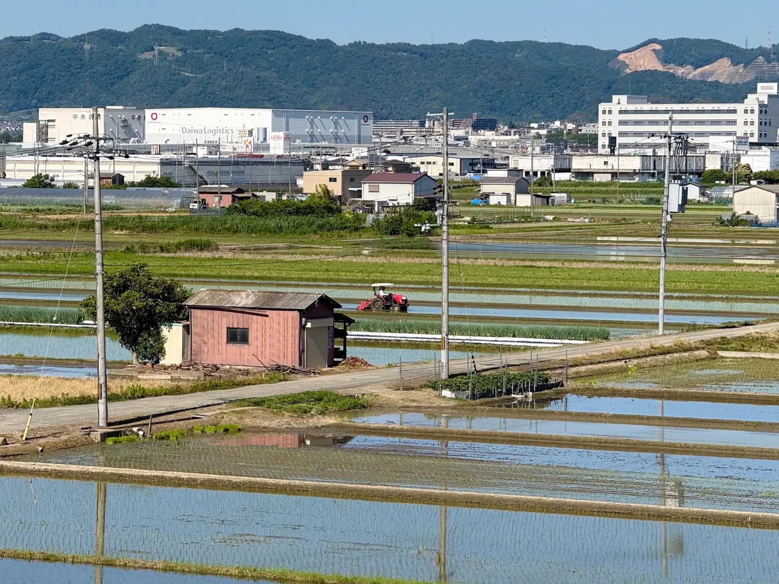 The farmland around the bridge: flooded, freshly planted rice paddies, a red tractor at work, and the logistics warehouses and hills of Yawata beyond.