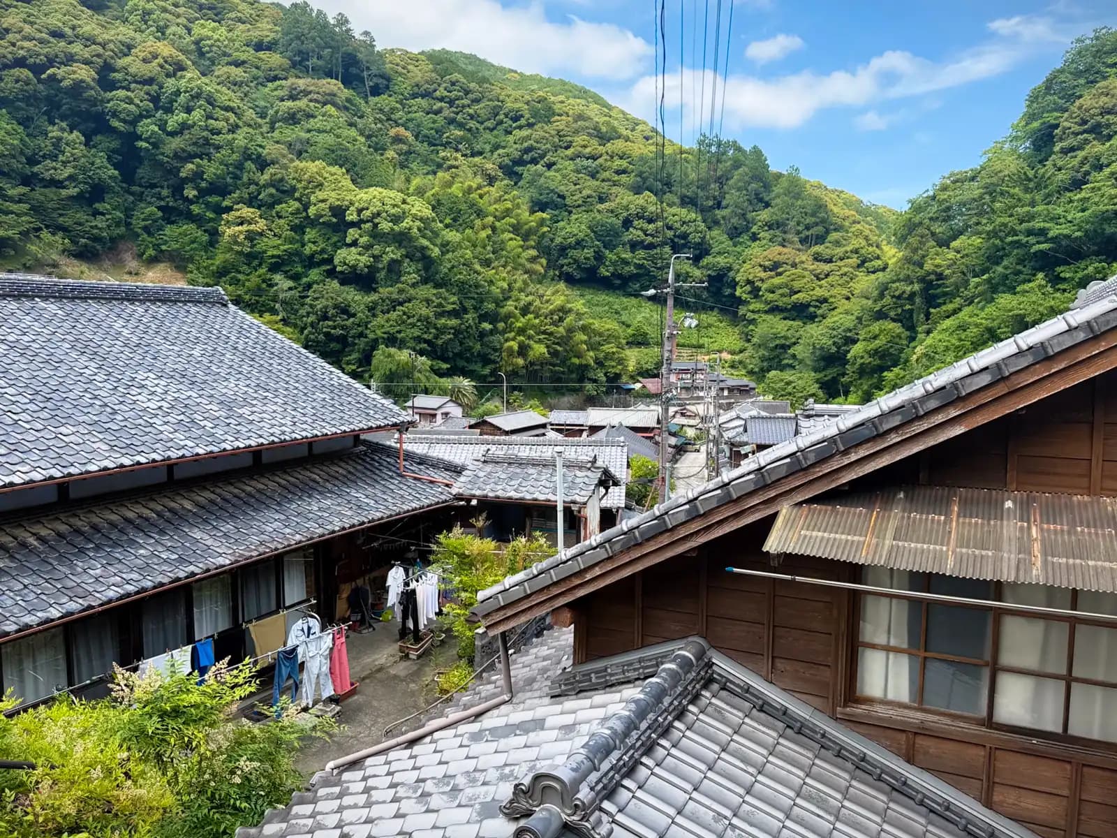 Looking out over the village rooftops from higher up. Tiled roofs, laundry lines, and the forested hill behind.