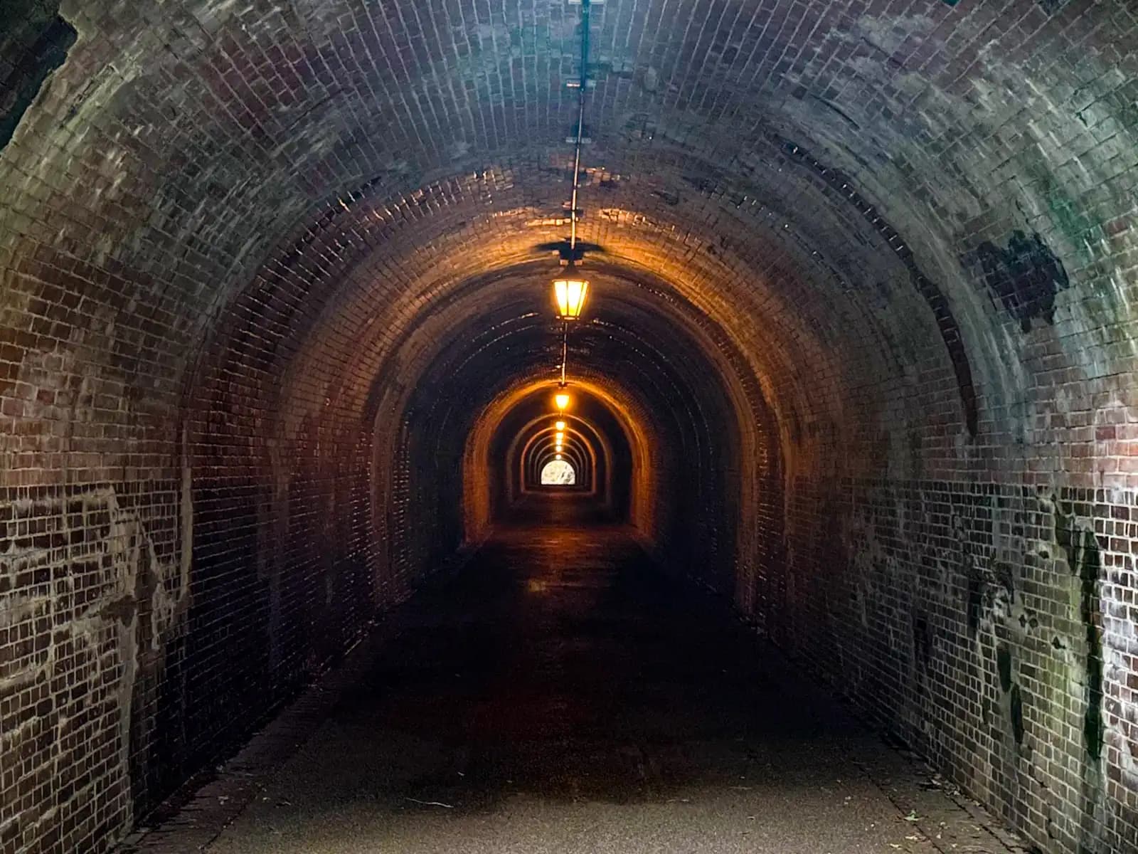 Inside the tunnel. The brick vault runs off into the dark, lit by a line of hanging lamps, with daylight at the far end.