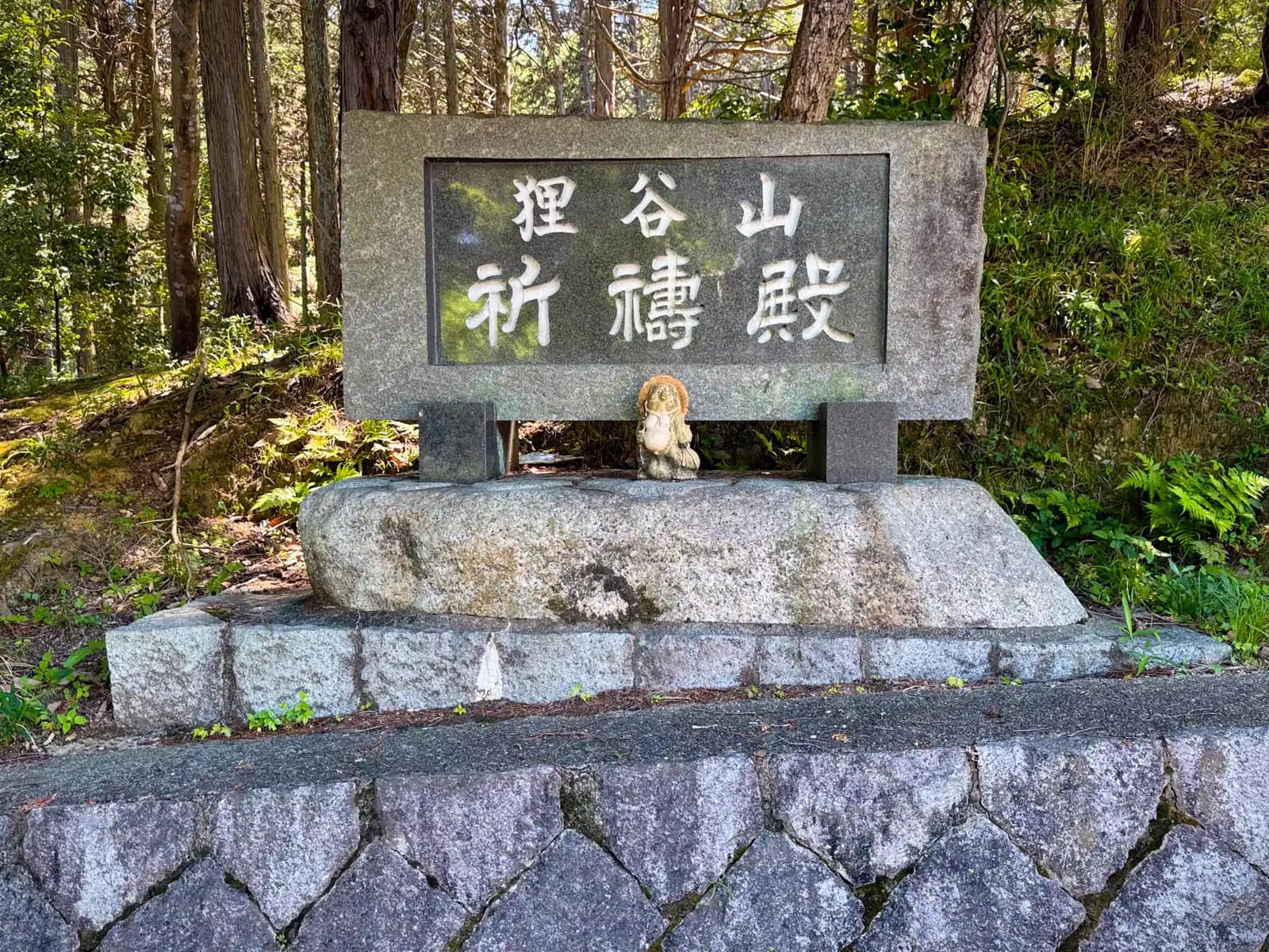 The carved stone marker for Tanukidani-san Fudō-in's prayer hall, a lone tanuki keeping watch beneath it in the cedar shade.
