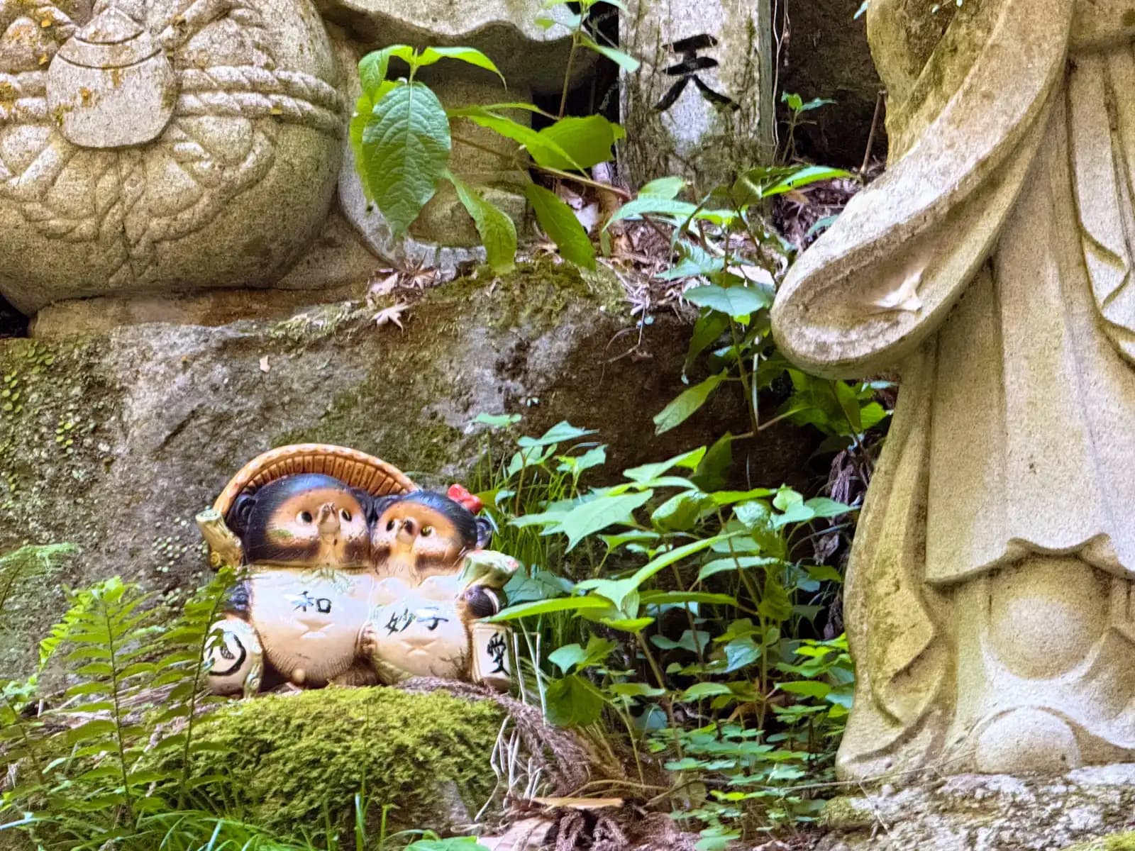 A pair of tanuki tucked at the foot of a robed stone deity, holding tablets that read harmony and love.