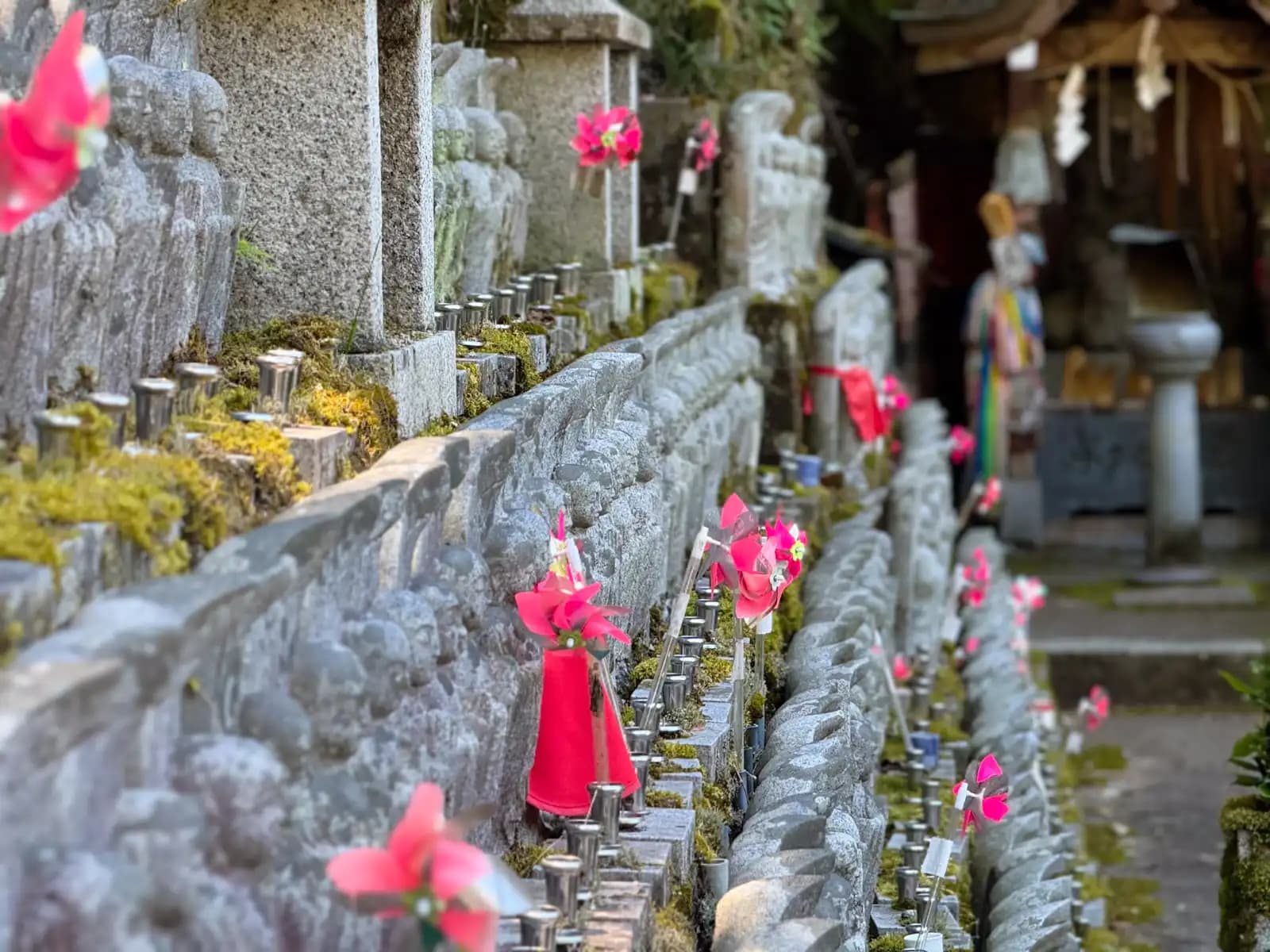 Rows of small stone figures line a mossy wall, bright pinwheels turning among them.
