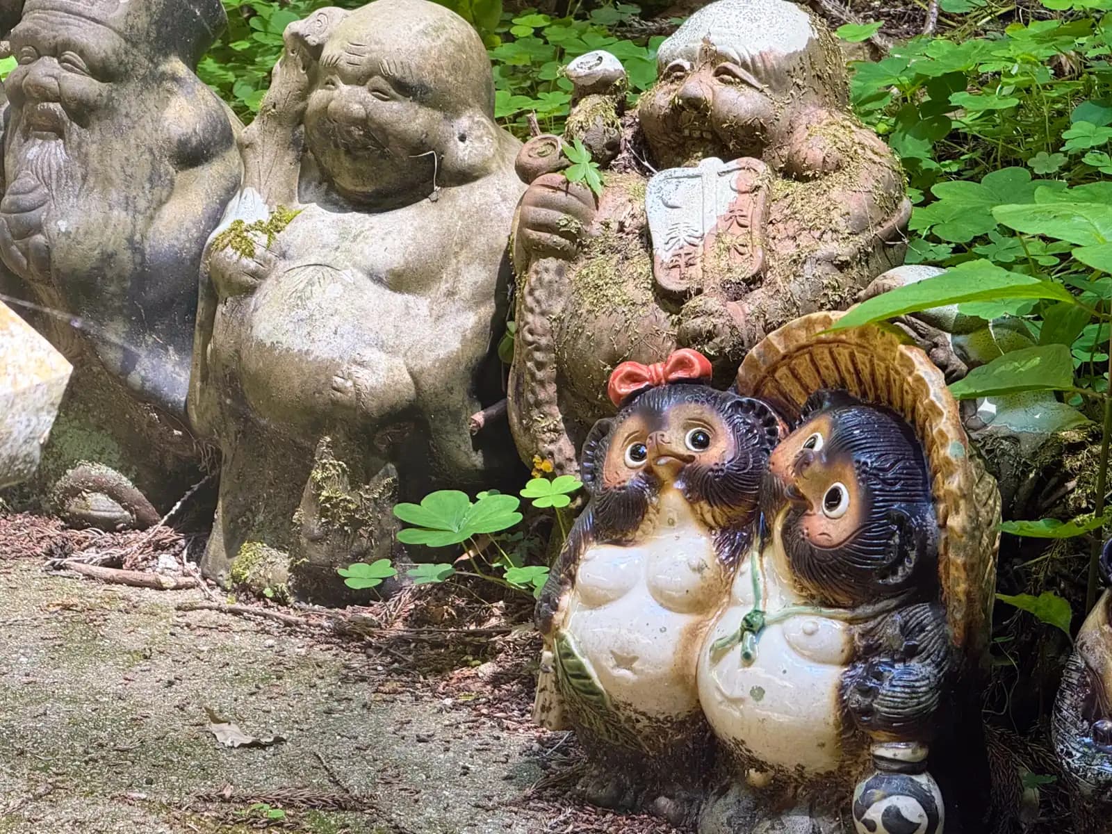 Mossy stone gods and, at their feet, a cheerful tanuki couple, one wearing a little red bow.
