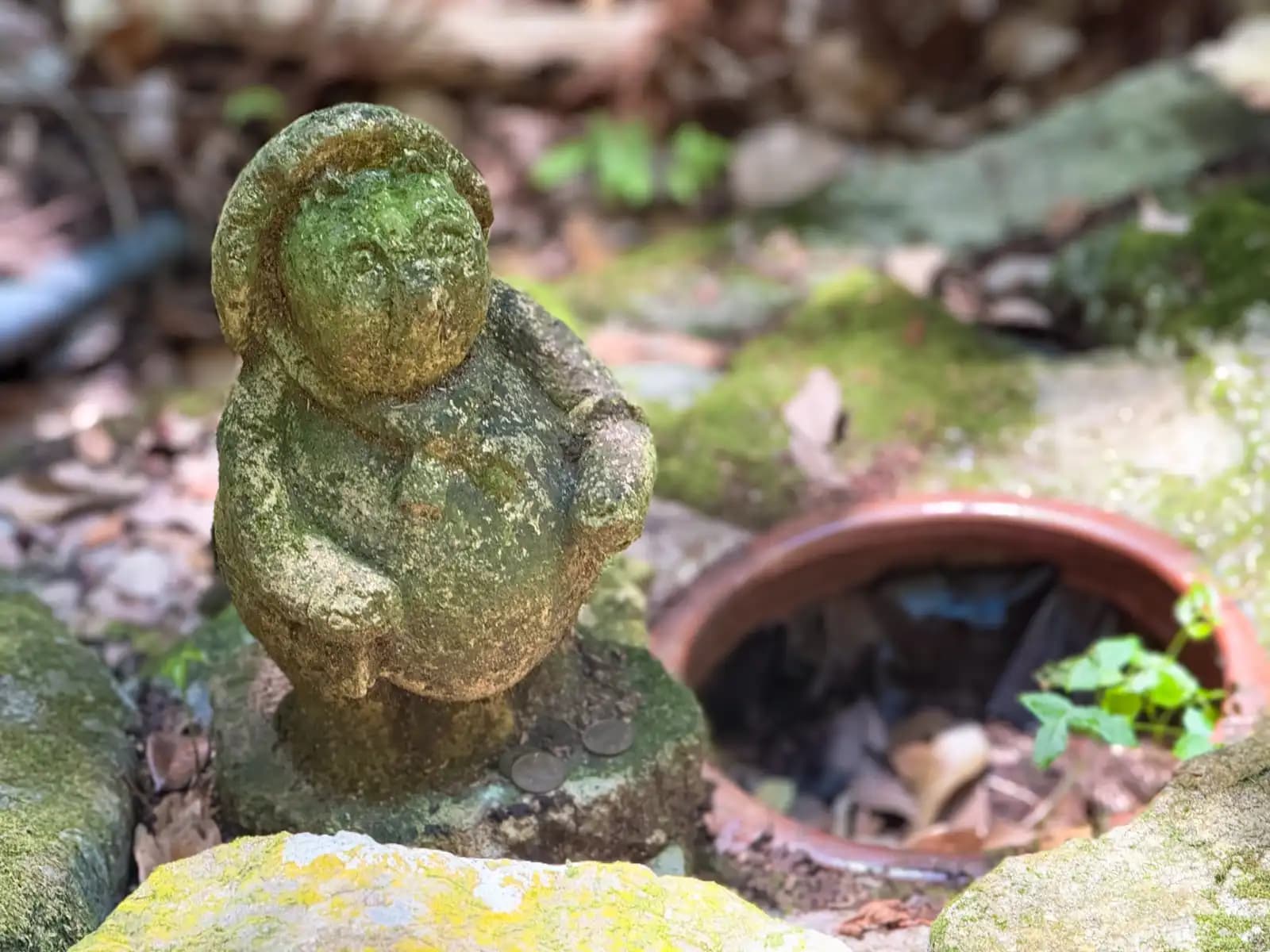 A tiny moss-covered figure, coins tossed at its base beside a half-buried offering pot.