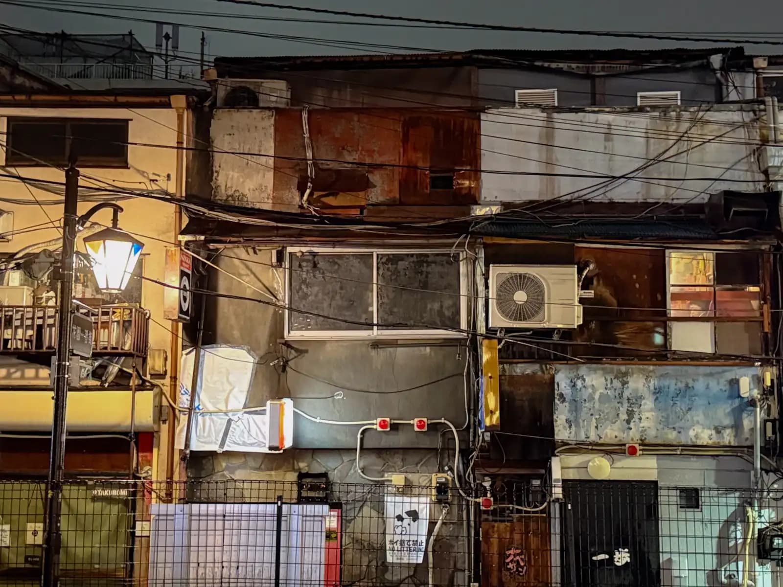 A row of old Golden Gai bars seen from the side of the alley: rusty metal siding, tangled wires and an old street lamp.