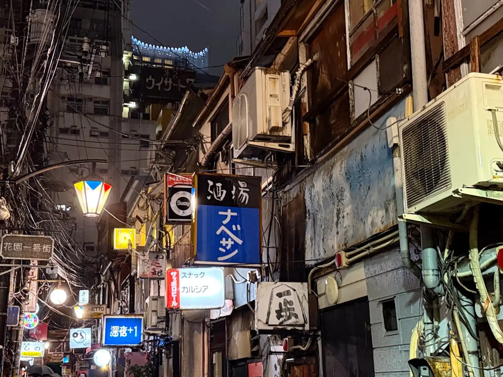 Signs stacked up along an alley at night, including Sakaba Nabesan, Snack Garcia and Shinya +1, with old air conditioners hanging off the walls.