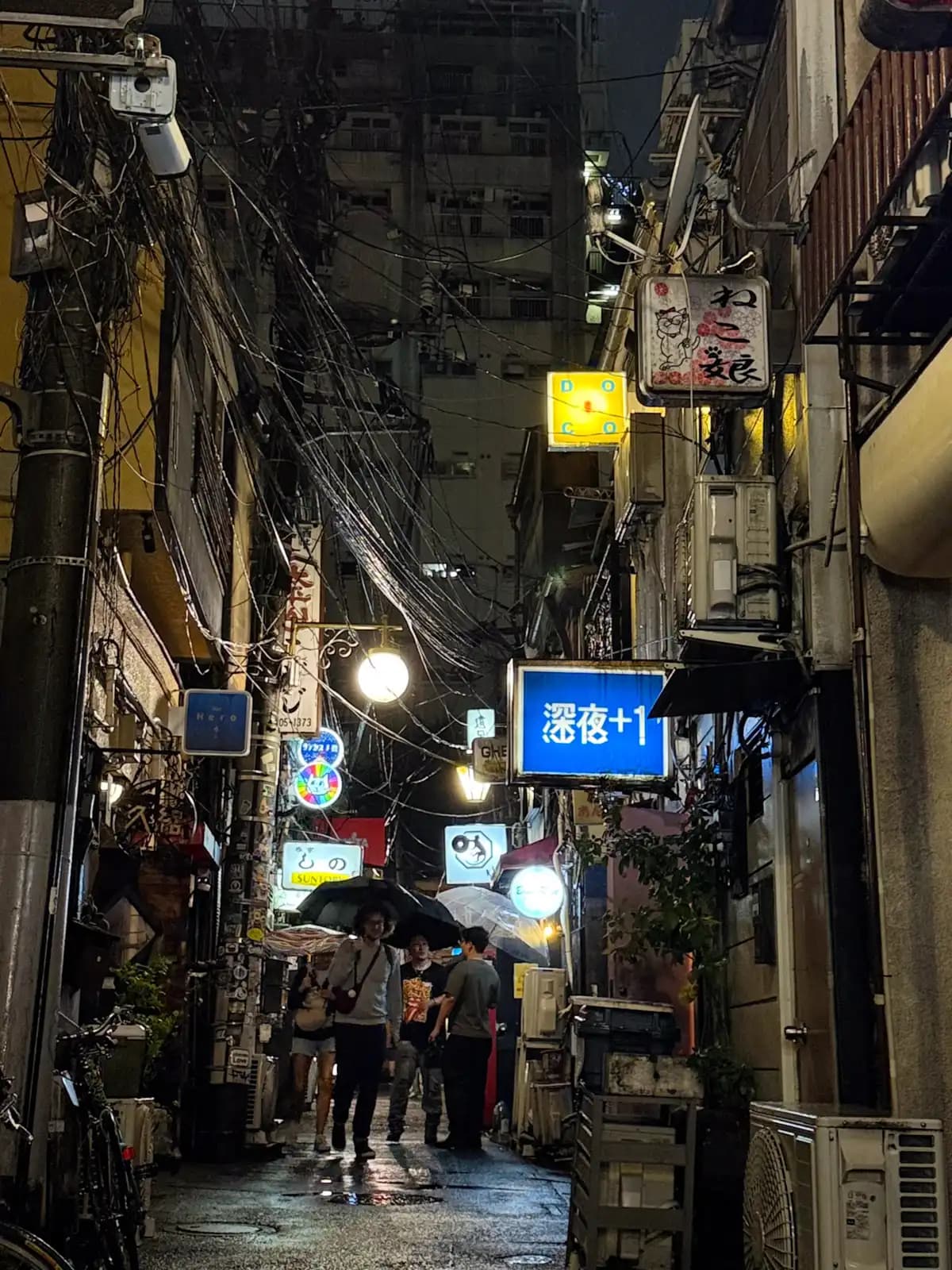 People with umbrellas walk down a wet alley on a rainy night, under a mess of power lines and the blue Shinya +1 sign.
