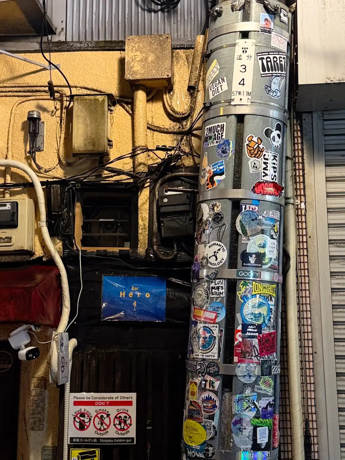 A pipe covered in stickers next to the door of Bar Hero, with a sign asking people not to be loud, smoke or drink outside.