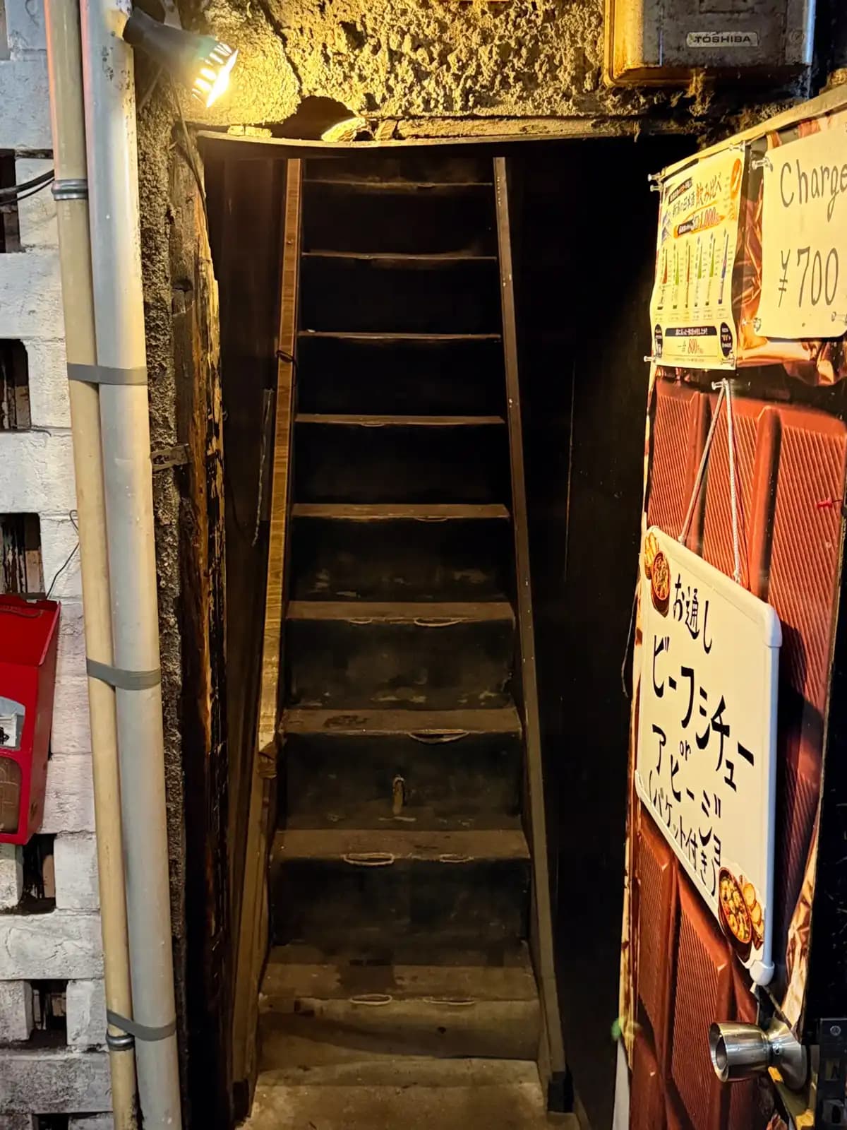 A very steep, narrow wooden staircase up to a second-floor bar. The sign says the cover charge is ¥700 and the snack is beef stew.