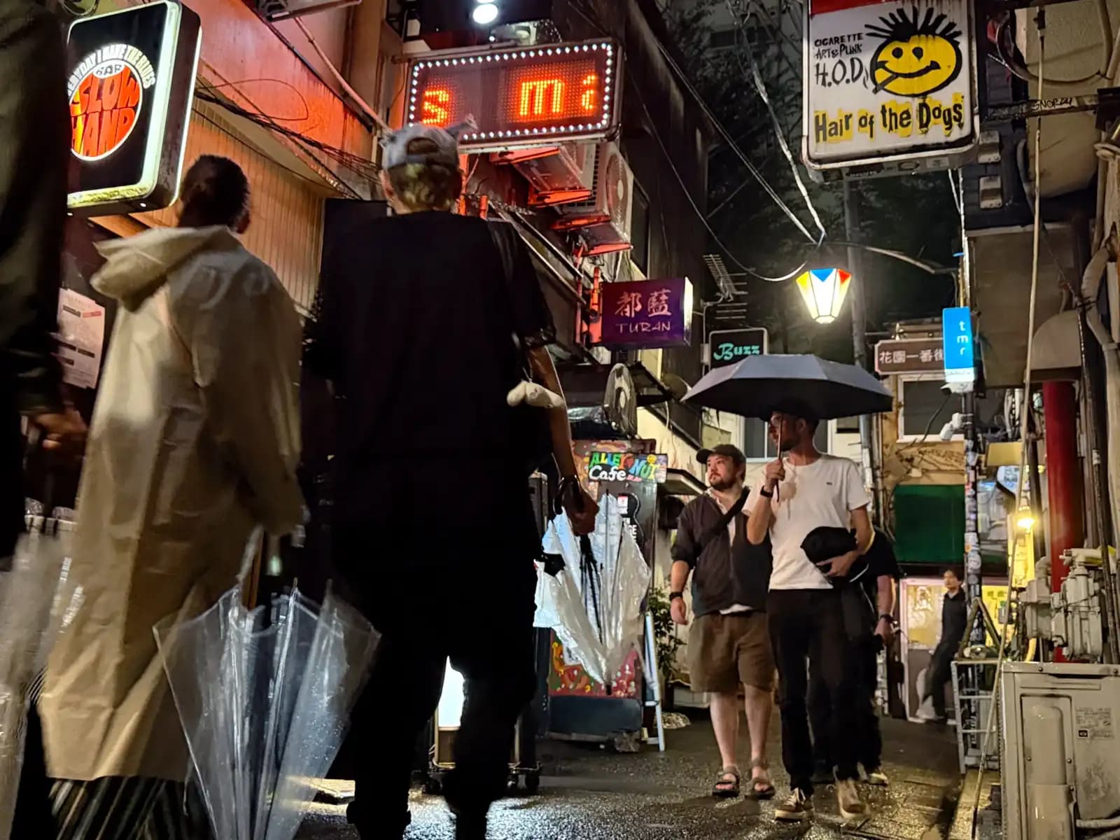People walking through the rain with clear umbrellas, past signs for Hair of the Dogs, Slow Hand and Turan.
